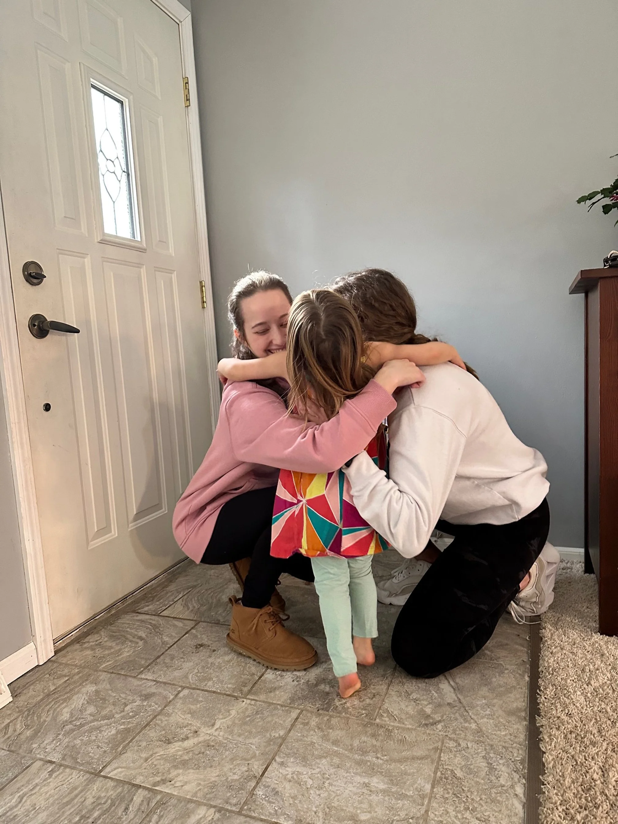 Three people, two adults and a child, hugging at the front door of a home, showing happiness as a MomsBloom Volunteer supports a postpartum family.