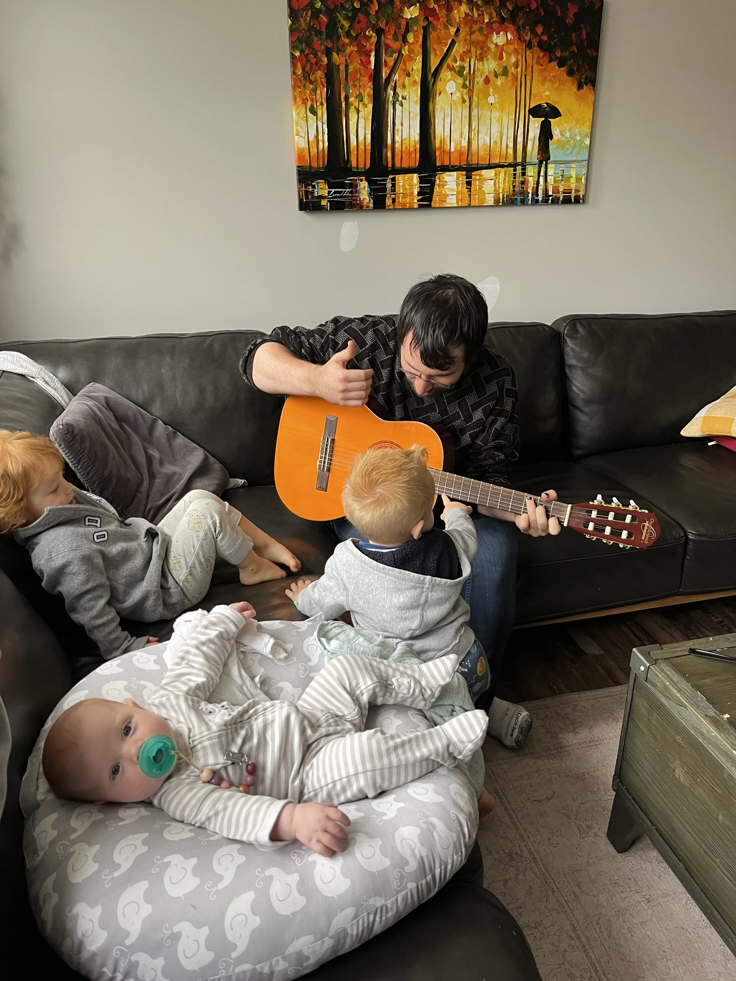 A man playing guitar with three children sitting around him on a couch and a baby on a cushioned seat. The room has a colorful autumn-themed painting on the wall.