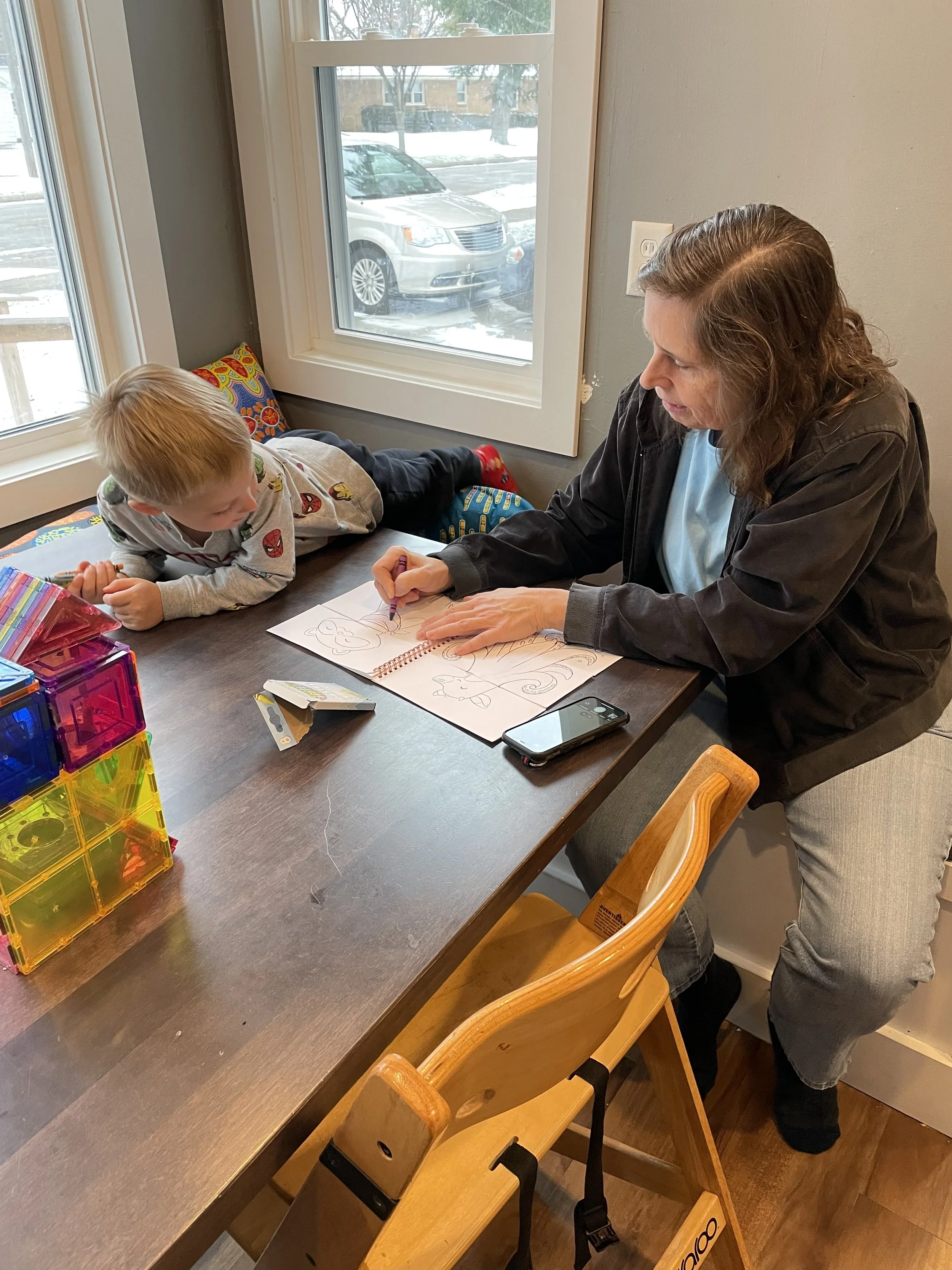 A woman is drawing in a sketchbook while a young boy watches from the table in a cozy indoor setting. There are colorful plastic storage blocks on the table and a window showing parked cars outside.