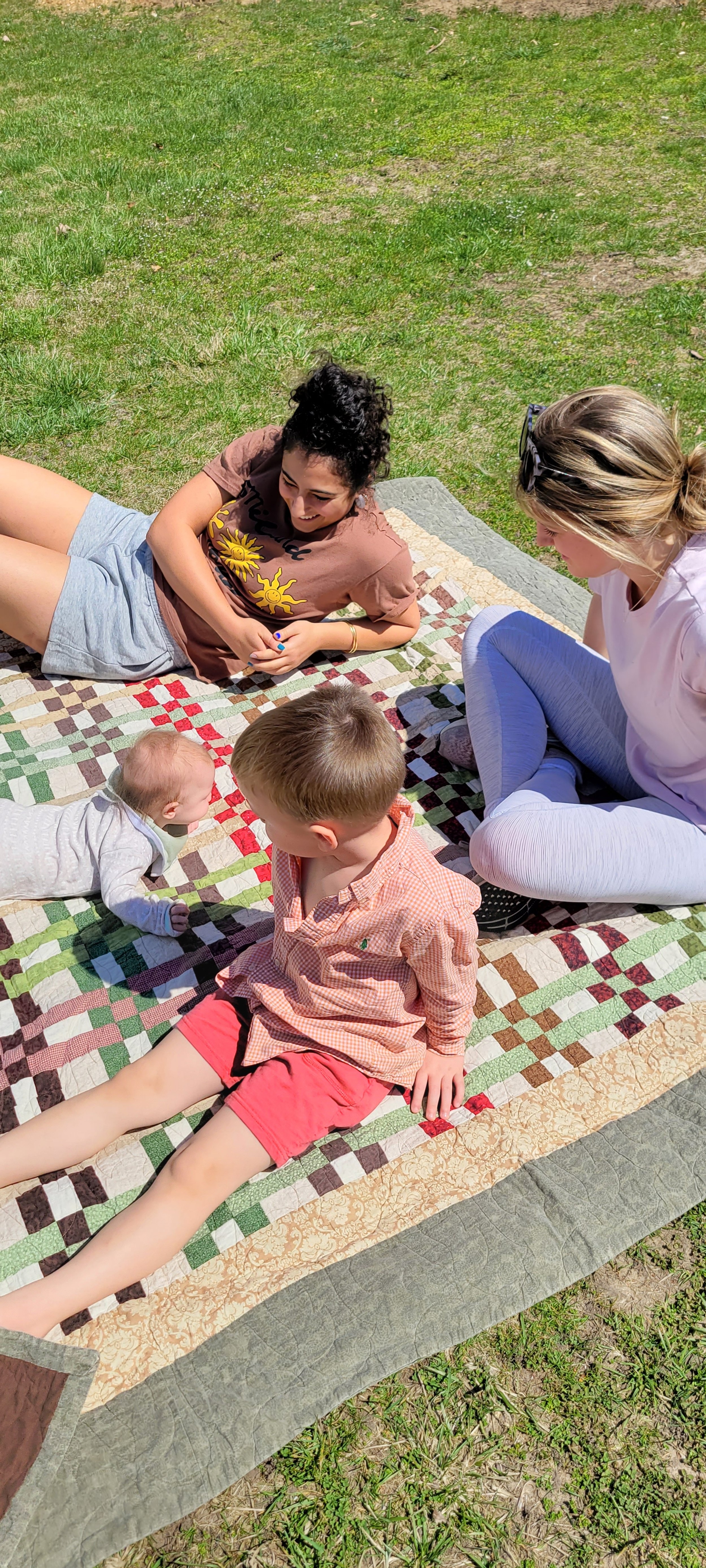 Four people, two adults and two children, lying on a patchwork quilt outdoors on grass, enjoying a sunny day.