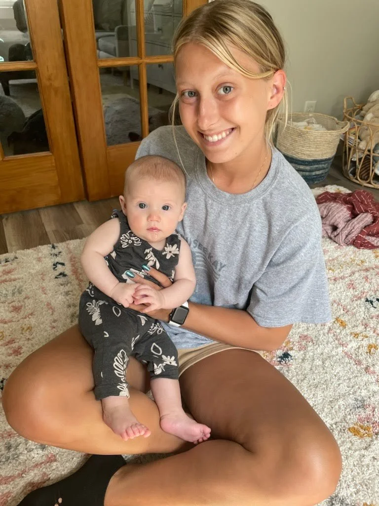 A smiling woman sitting cross-legged on a patterned rug, holding a baby on her lap, with a wooden door and glass panels in the background.
