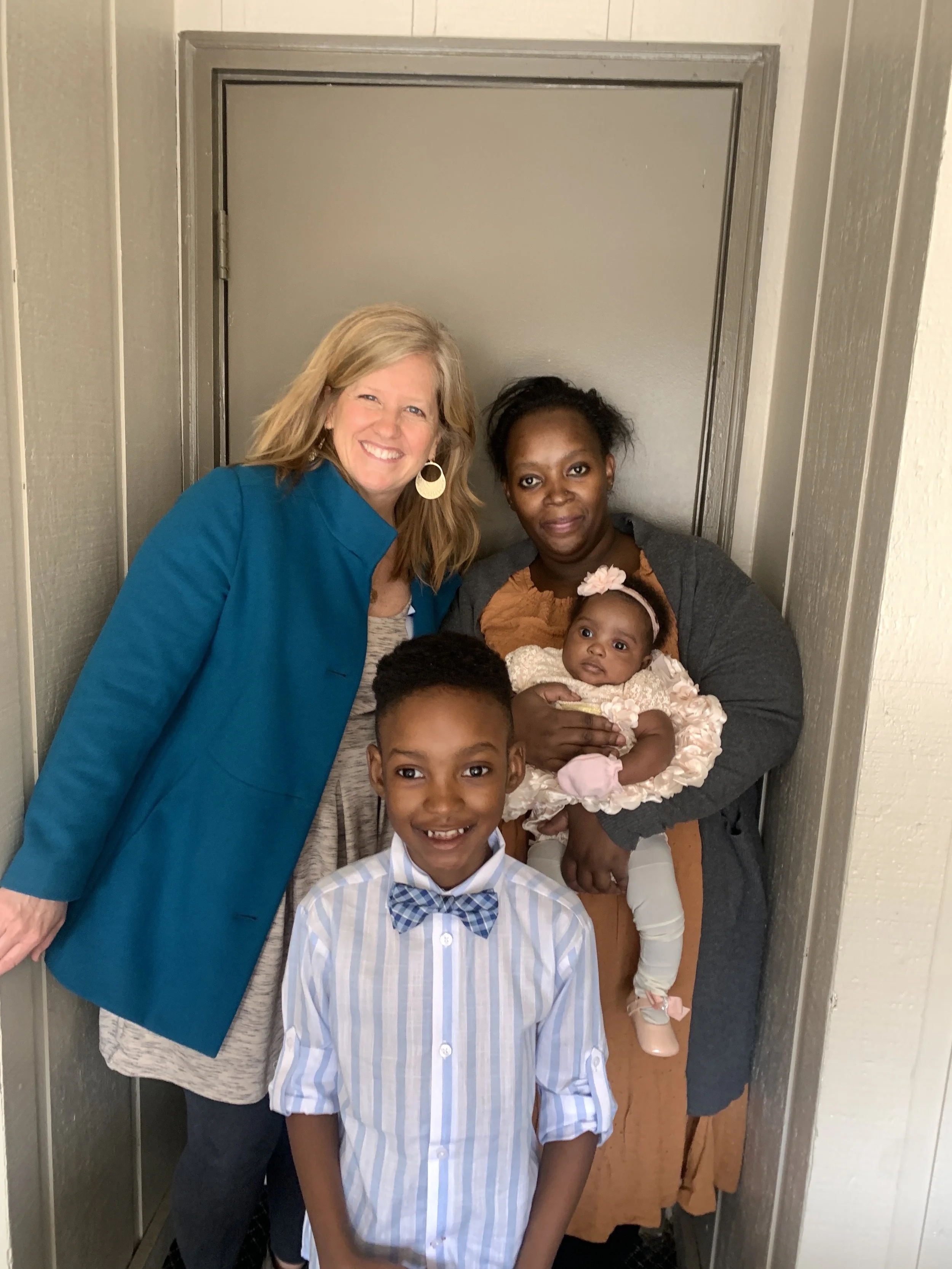 Four people inside an elevator: a smiling woman with blonde hair in a blue coat on the left, an African American woman with dark hair and a gray sweater holding a baby girl in a pink dress on the right, and a young boy with short dark hair, wearing a