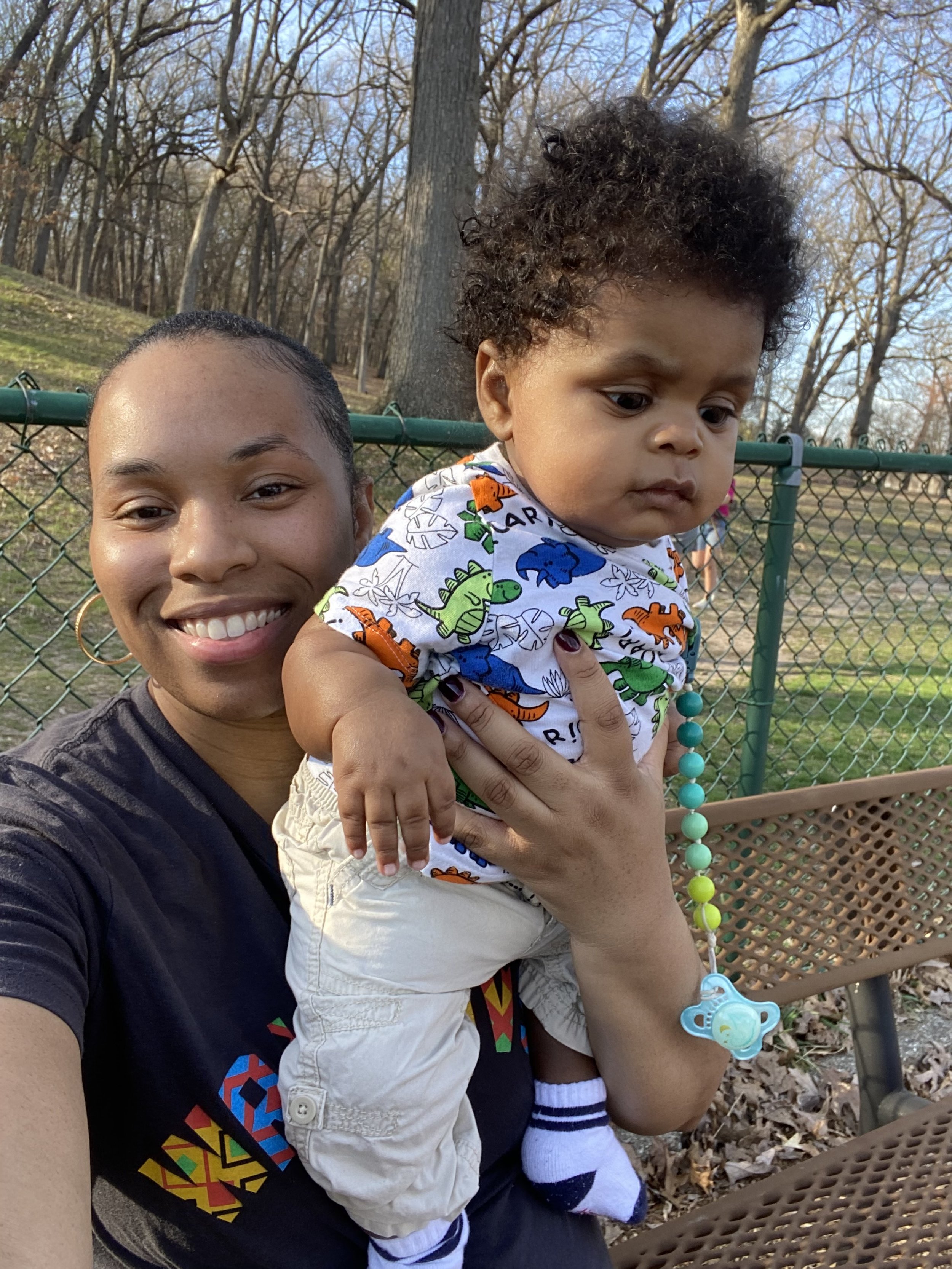 A woman with dark hair and earrings holding a young child on a park bench. The woman is smiling at the camera, while the child has curly hair and looks away. The background shows a chain-link fence and trees.