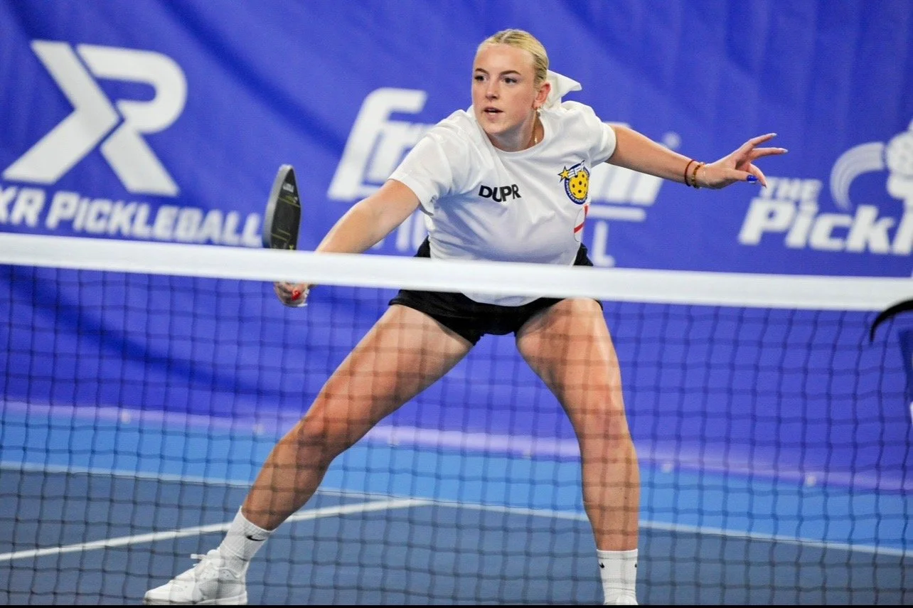 A young woman playing pickleball, preparing to hit the ball at the net, wearing a white shirt with the name 'DUPR' and a logo, black shorts, and white athletic shoes, on a blue court with a blue backdrop.