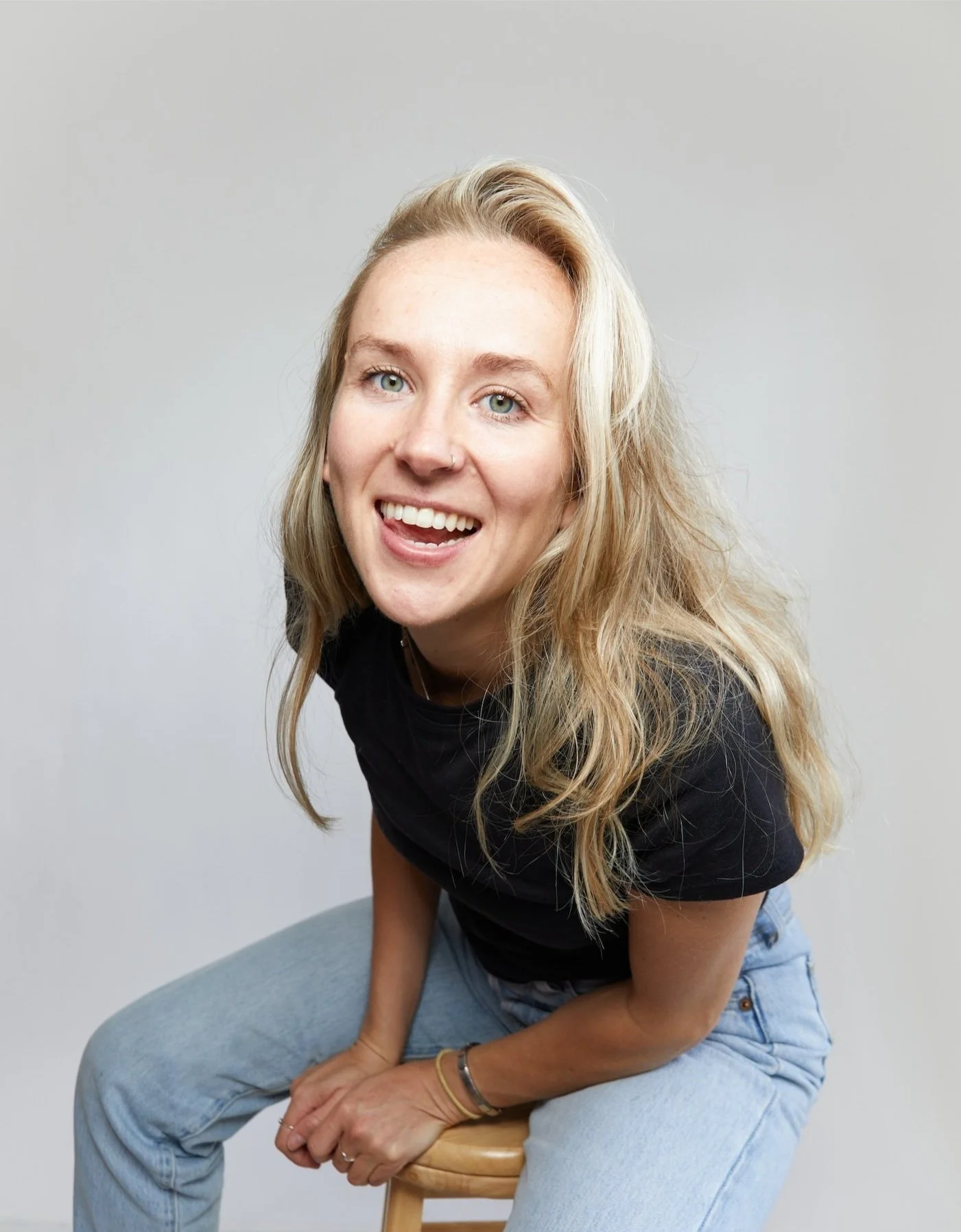 A woman with blonde hair, green eyes, and a nose piercing smiling and sitting on a stool against a plain gray background.