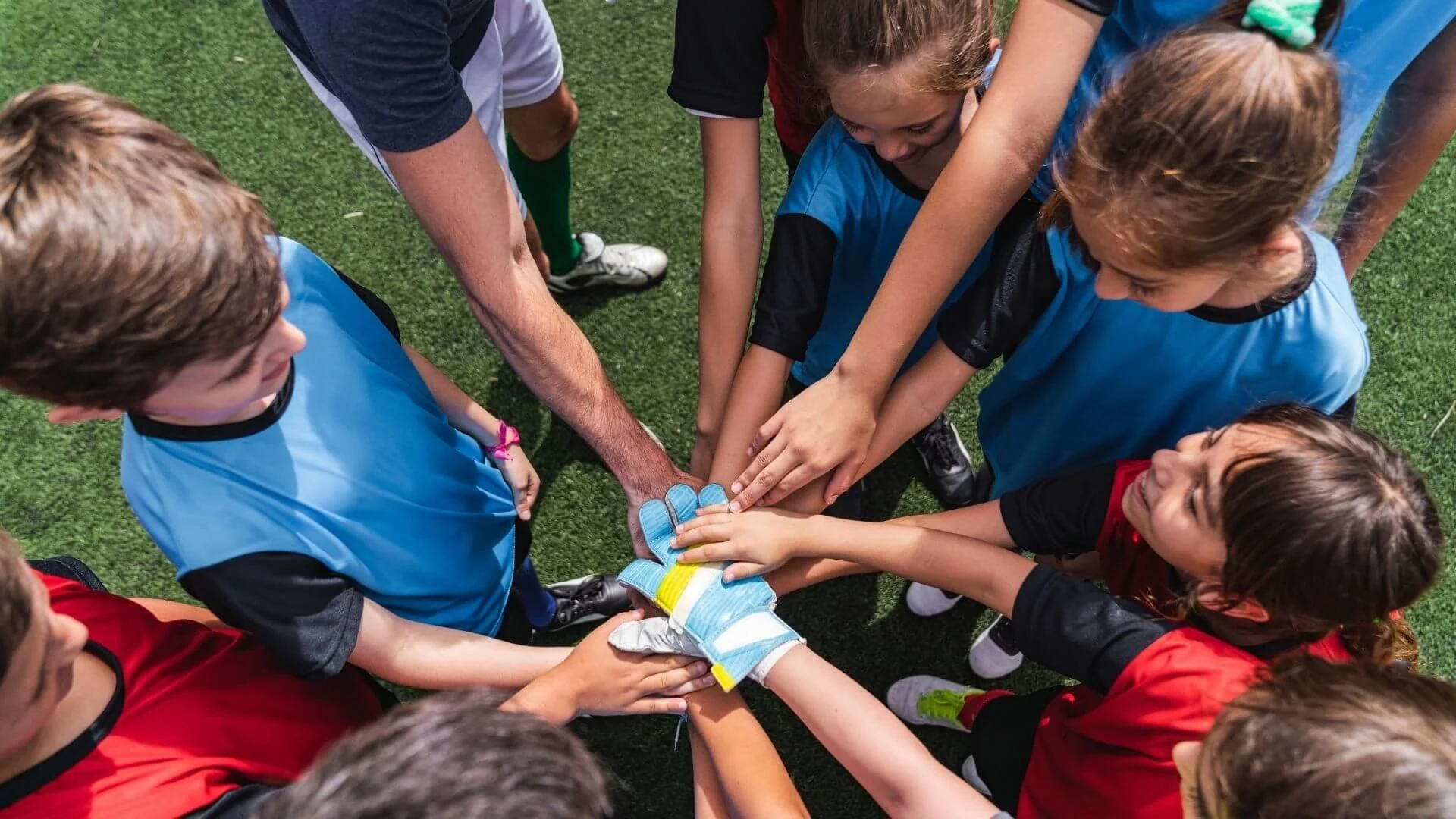 Group of young soccer players in colorful uniforms putting their hands together in the center on a green turf field.