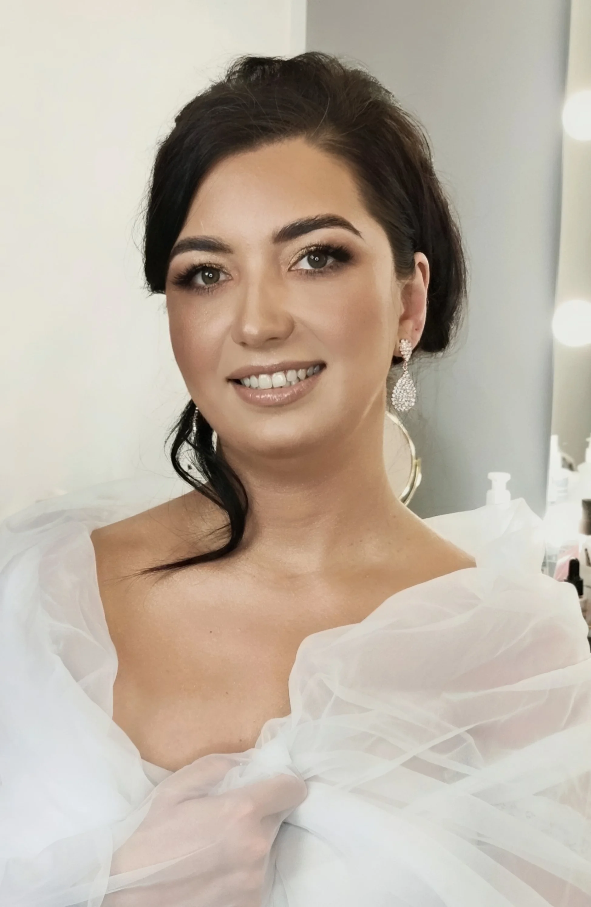 Close-up of a woman with dark hair and earrings, smiling, wearing a voluminous white dress with sheer fabric details, in a well-lit room.