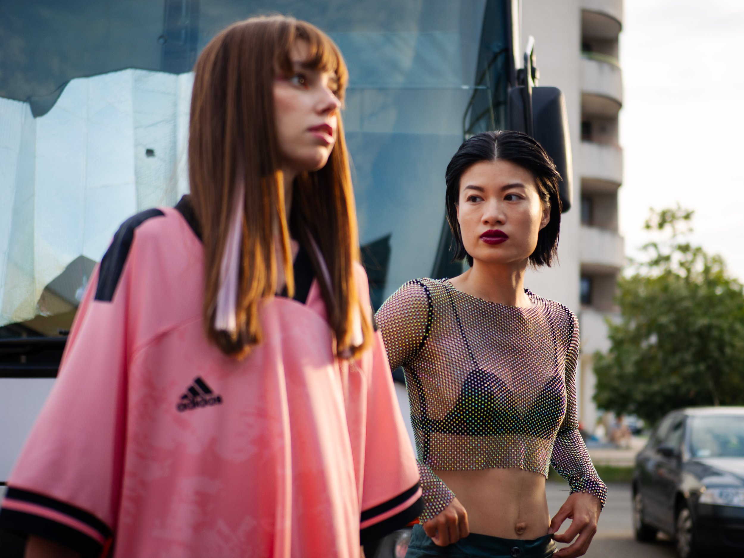 Two women standing outdoors near a large vehicle, with buildings and trees in the background. One woman has long brown hair and is wearing a pink Adidas jacket, and the other woman has short black hair and is wearing a sparkly mesh top with black bra