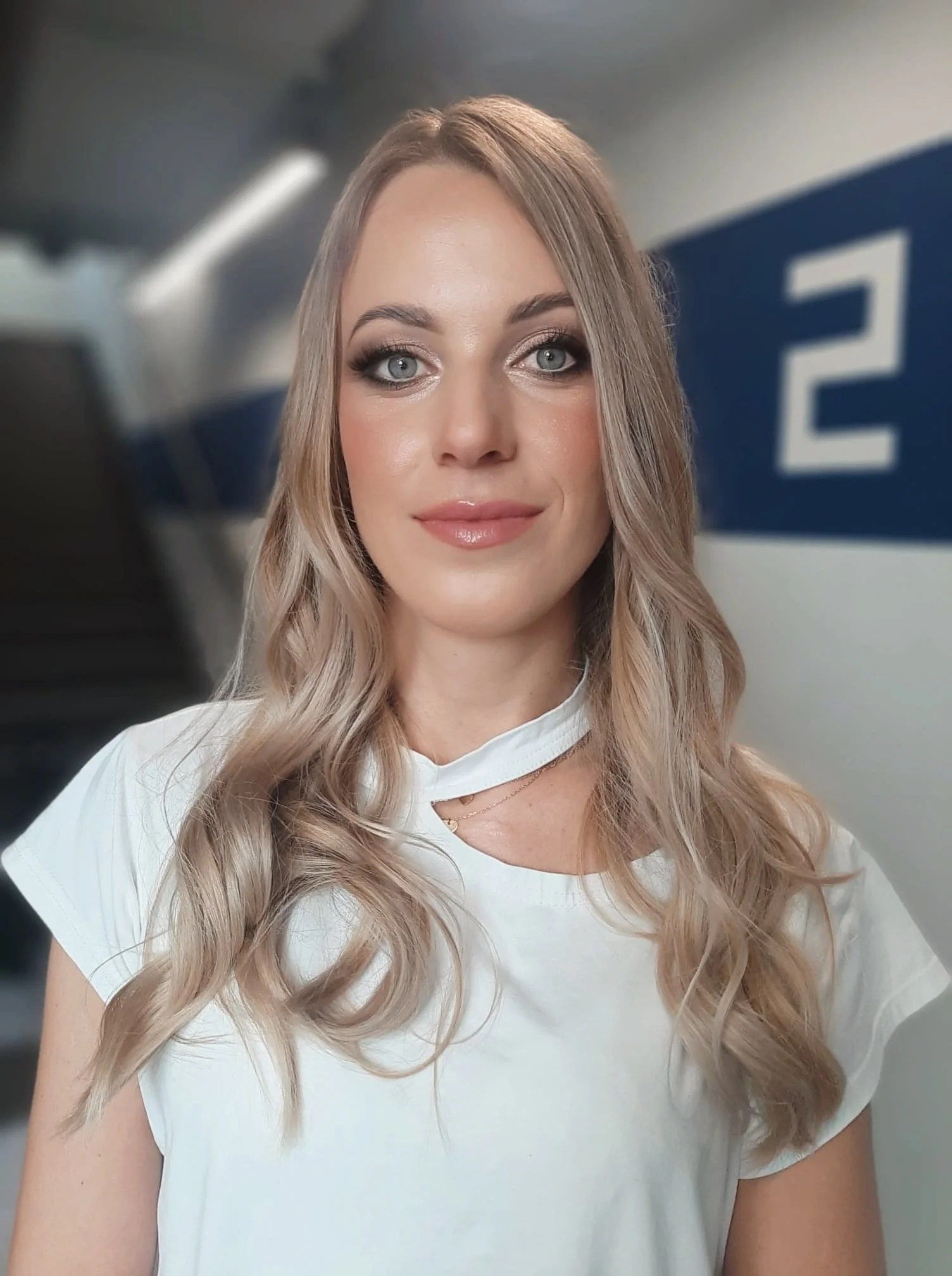 A young woman with long, wavy blonde hair and blue eyes, wearing a white top, standing indoors with a blue and white sign in the background.