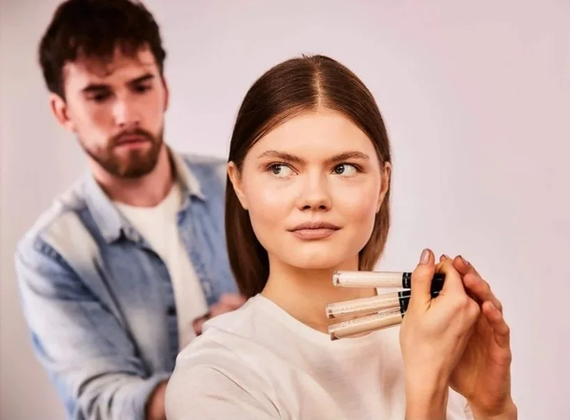 A woman holds multiple makeup markers while sitting in a beauty salon, with a makeup artist behind her.