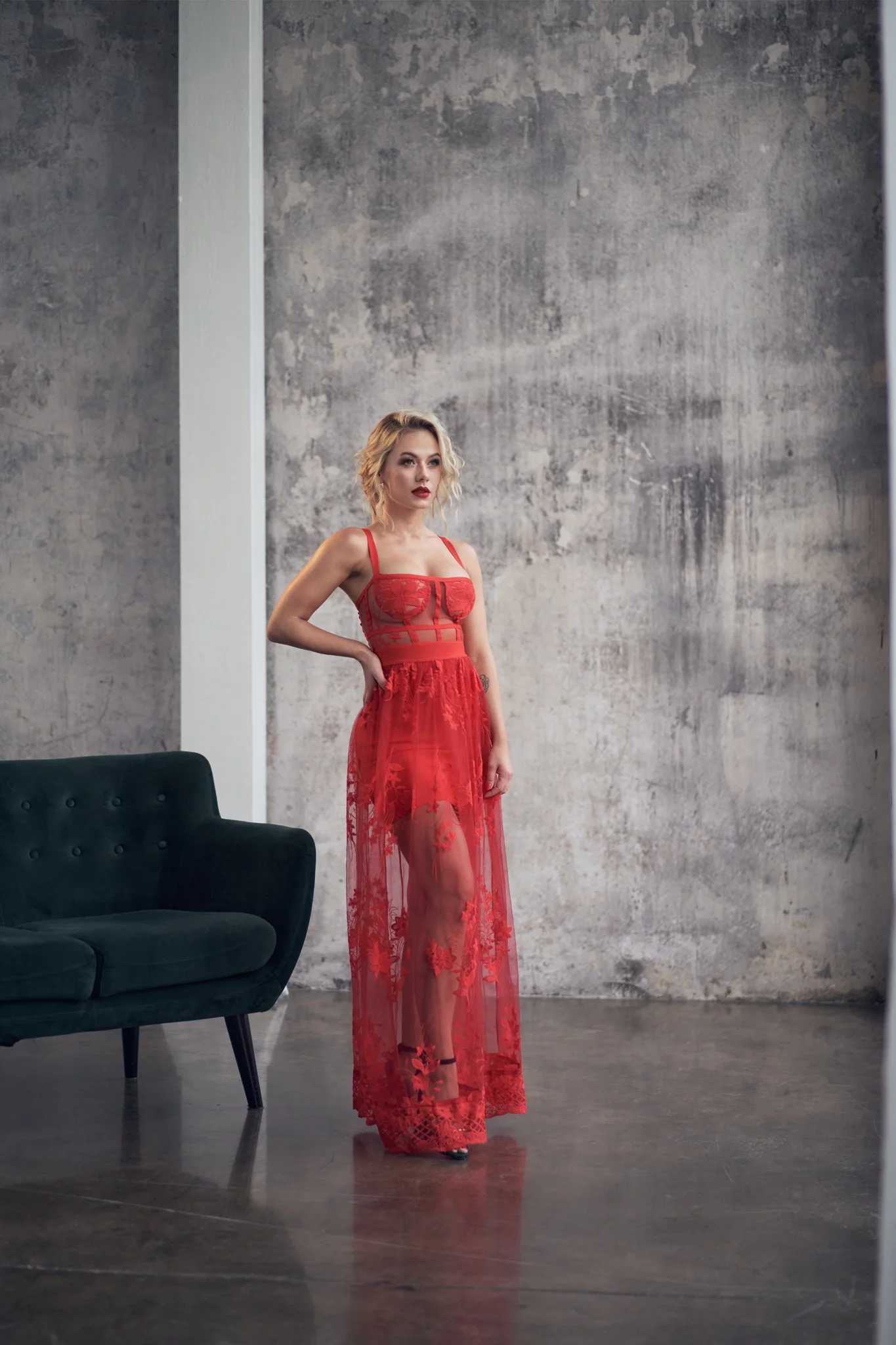 A woman with blonde curly hair and red lipstick stands in an industrial-style room wearing a red sheer lace dress, with one hand on her hip next to a dark green velvet sofa, against a textured gray wall.