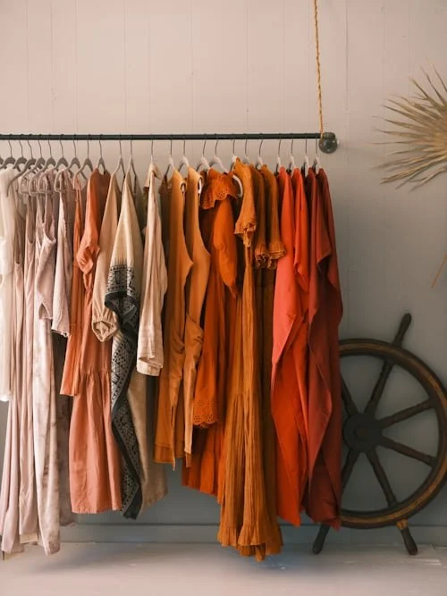A row of hanging women's dresses in neutral, white, peach, and rust colors on a black clothing rod, with a wheel and a leafy plant in the background.