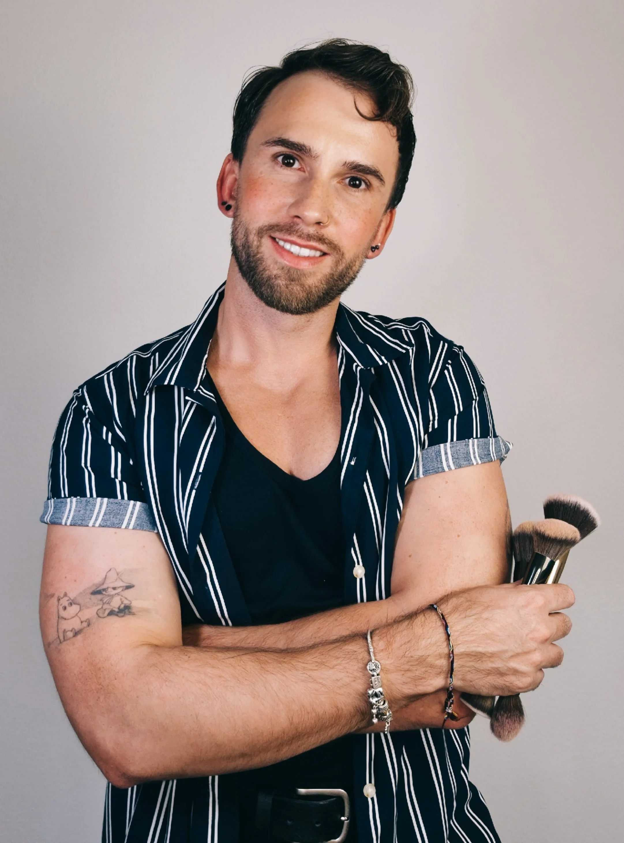 A smiling man with dark hair and beard, wearing a striped short-sleeve shirt and holding makeup brushes, standing in front of a neutral background.