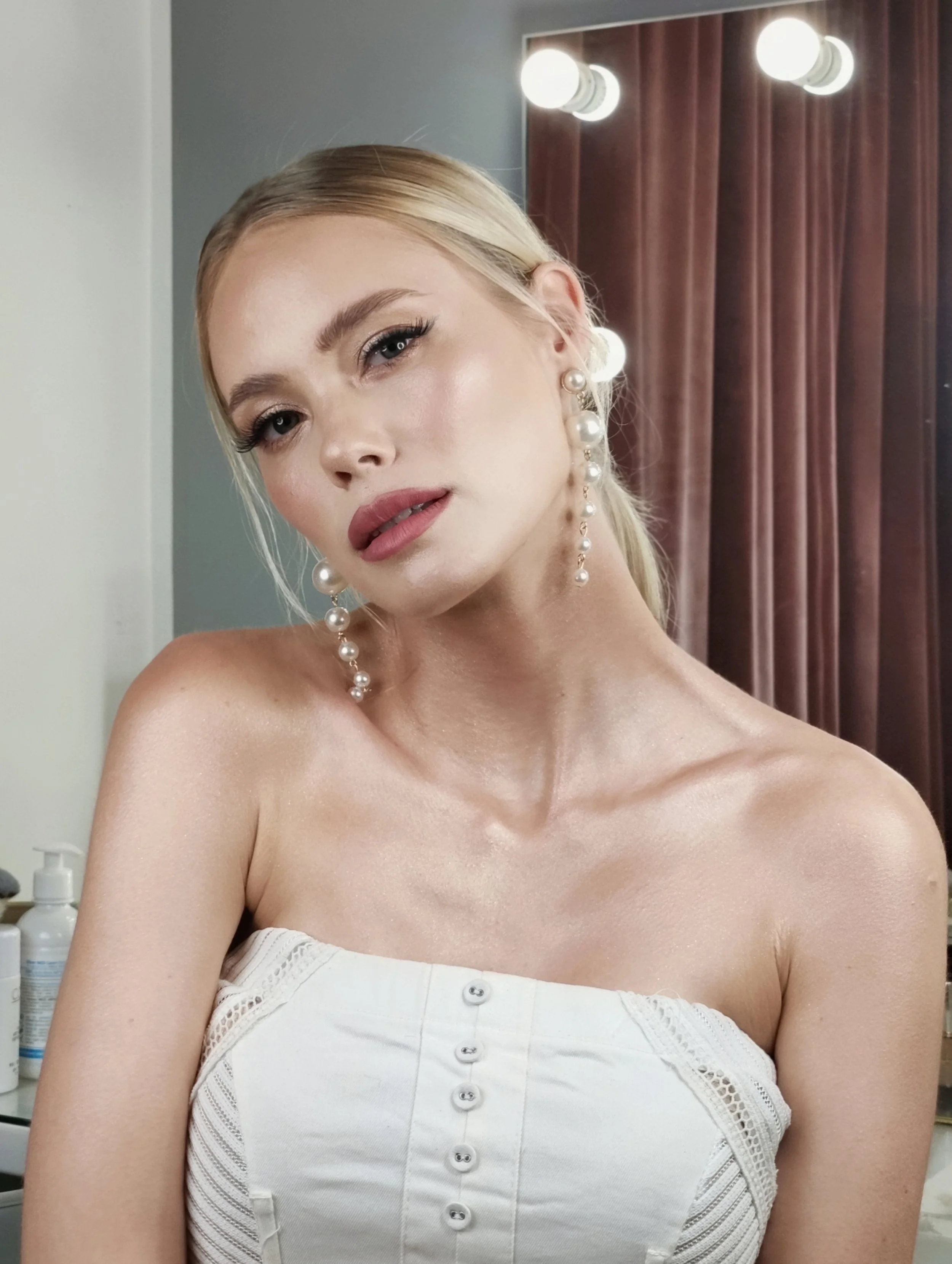A young woman with blonde hair styled in a ponytail, wearing pearl earrings and a strapless white top with buttons, looking into the camera in a room with brown curtains and bright lights in the background.