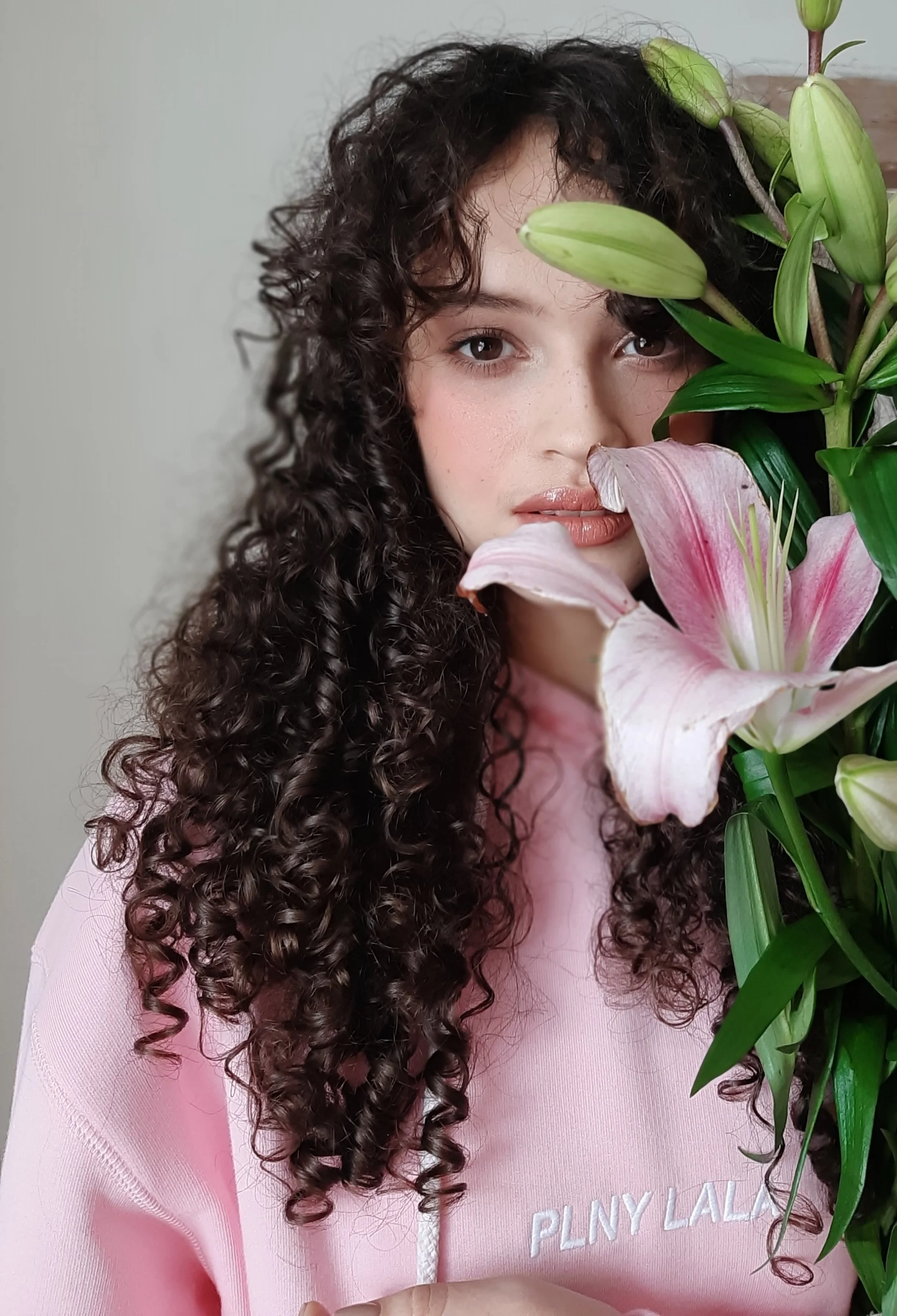 A young woman with curly dark hair and wearing a pink sweatshirt with white text, standing behind pink lilies and green foliage, looking at the camera.