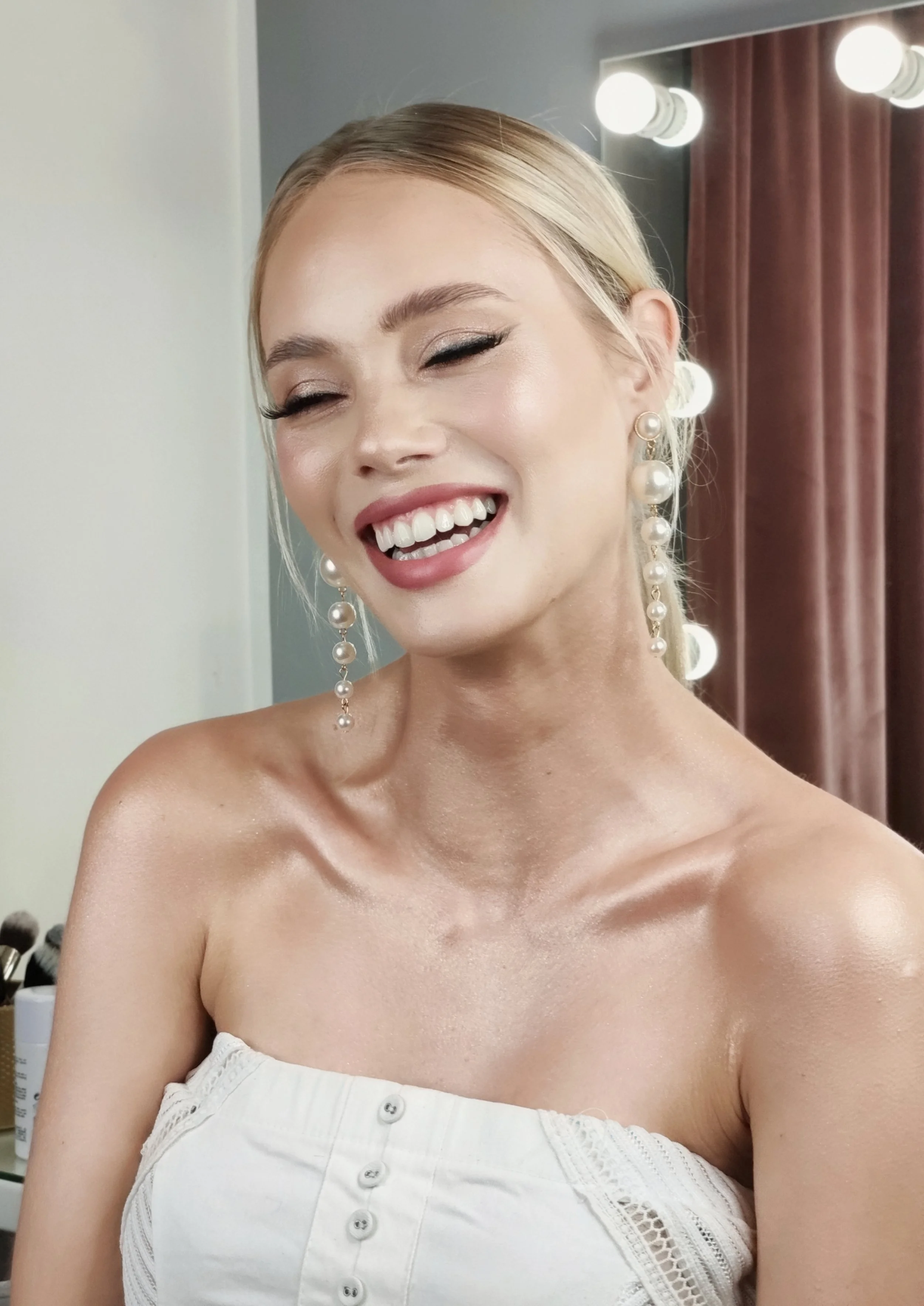 A woman with blonde hair, wearing pearl earrings and a white strapless top, smiling with her eyes closed in a dressing room with bright lights and brown curtains.
