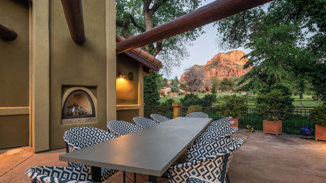 Outdoor patio with a long dining table and patterned chairs, fireplace on the wall, surrounded by trees and a clear view of red rock formations in the background.