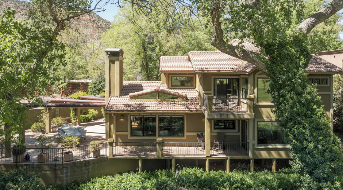 A two-story house with tan exterior and red-tiled roof, surrounded by trees and greenery, featuring a balcony and patio area.