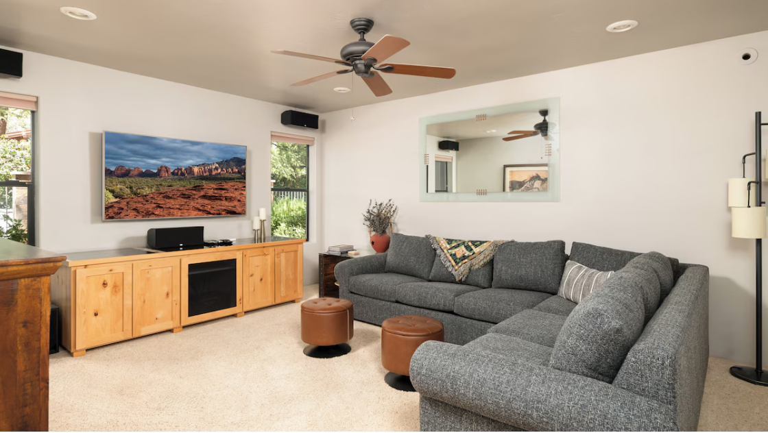 Modern living room with a large gray sectional sofa and decorative pillows, two brown ottomans, a wooden media console with a flat-screen TV, various decor pieces, and a ceiling fan.