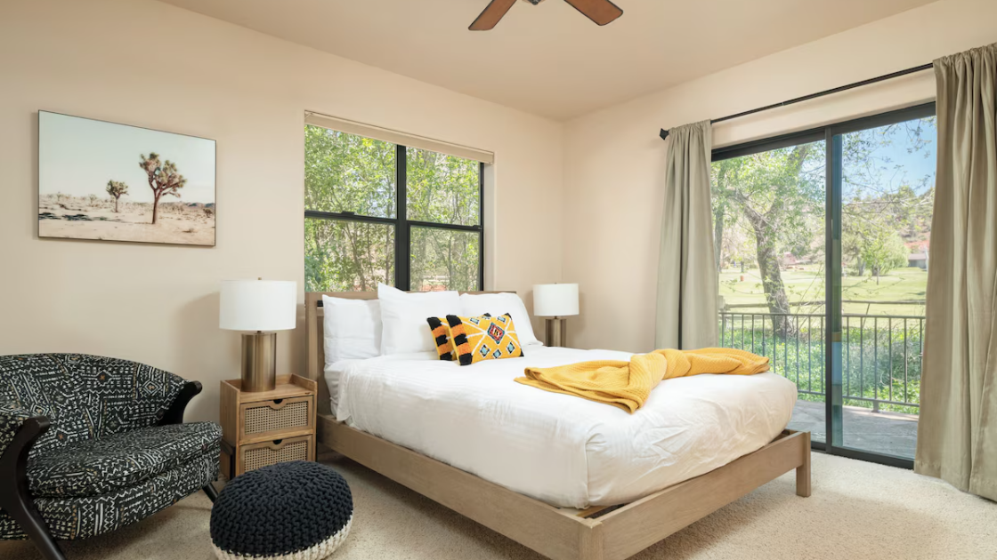 Bright bedroom with a bed, yellow blanket, decorative pillows, wooden nightstands with lamps, an armchair, round knitted ottoman, abstract wall art, large windows, and sliding glass door with natural light and outdoor view.