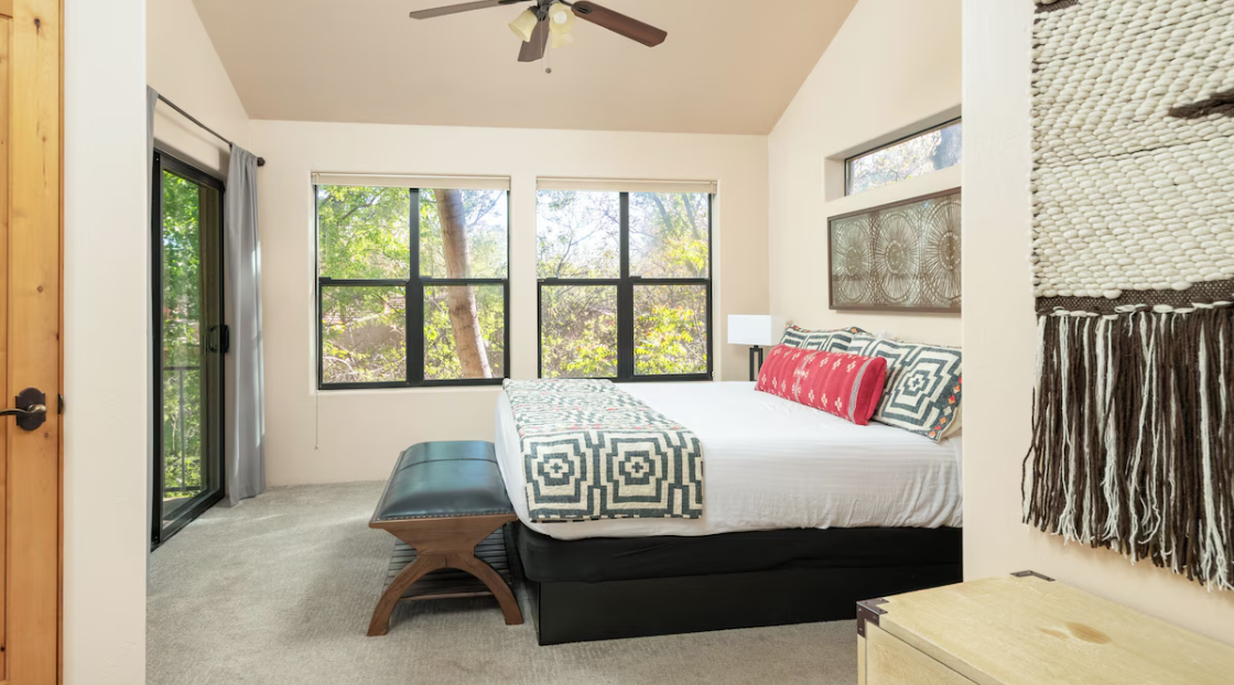 Modern bedroom with large windows, a bed with patterned pillows and blanket, a ceiling fan, wall art, and a sliding glass door.