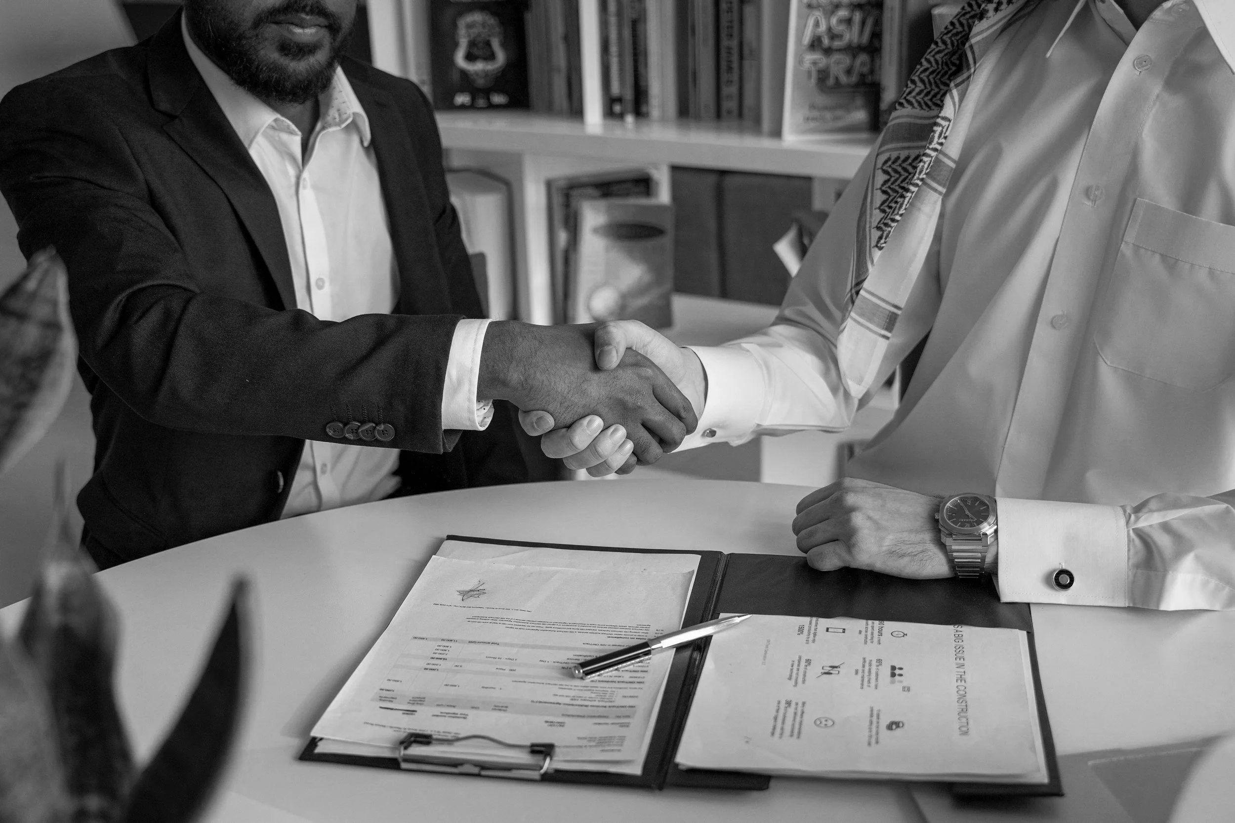Two men shaking hands across a table with documents and a pen, in an office setting.