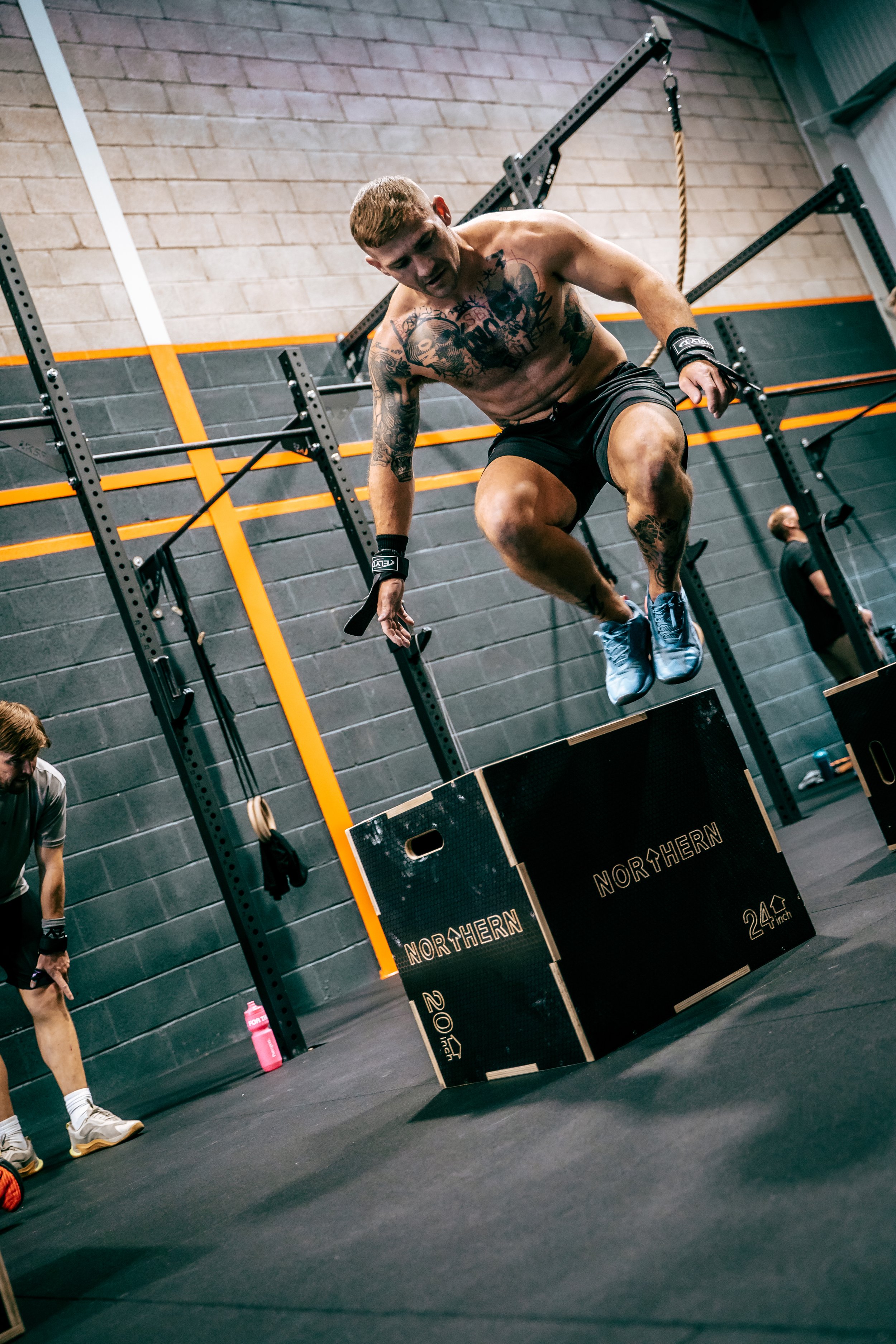A shirtless man with tattoos and wearing black shorts is jumping onto a box during a workout in a gym. The gym has a black and gray wall with orange accents, and exercise equipment is in the background. Other people are visible in the gym, including a person in the bottom left corner holding a water bottle.