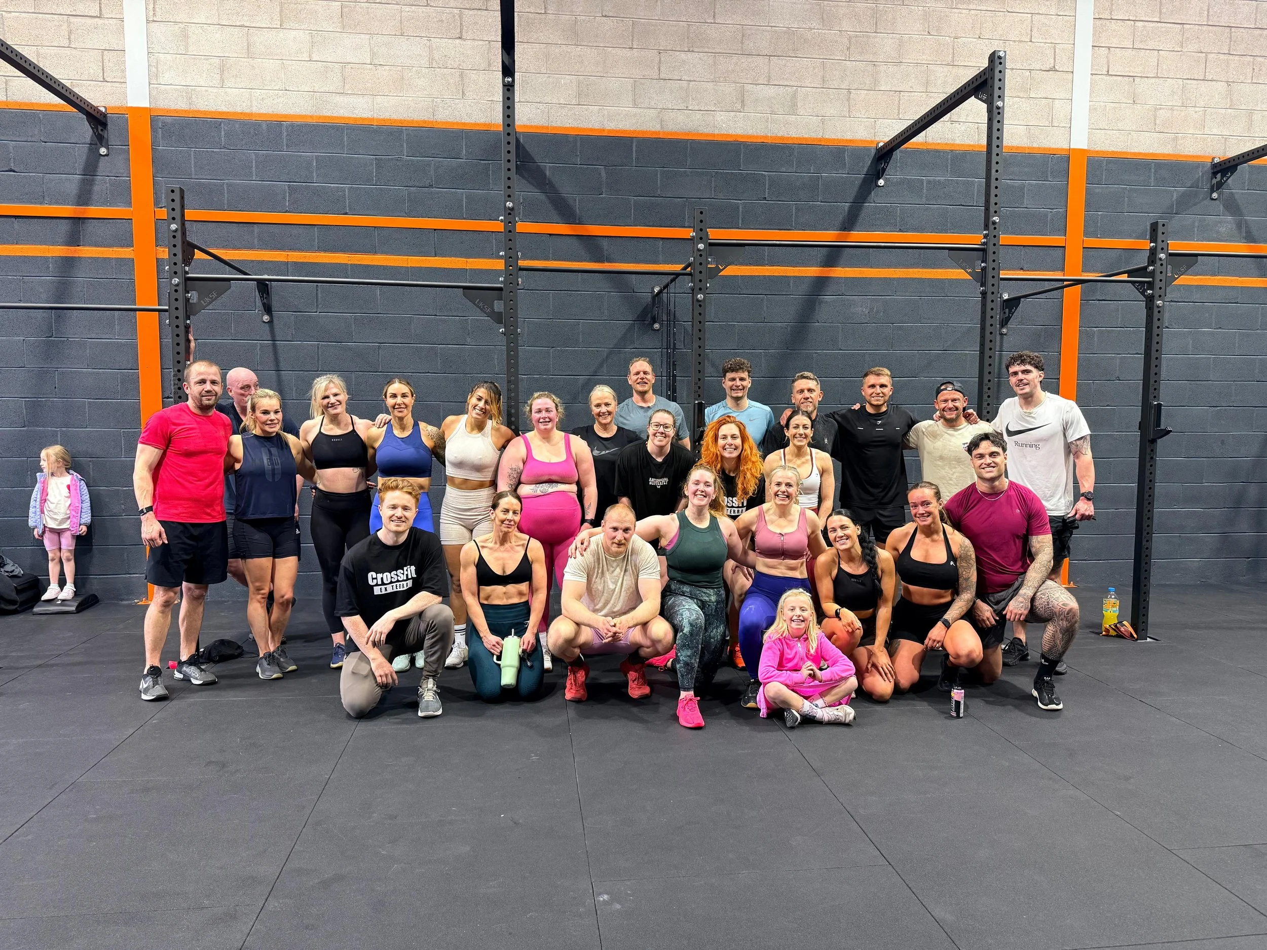 Group of diverse people in workout attire smiling at the camera inside a gym, with black and orange gym racks in the background.