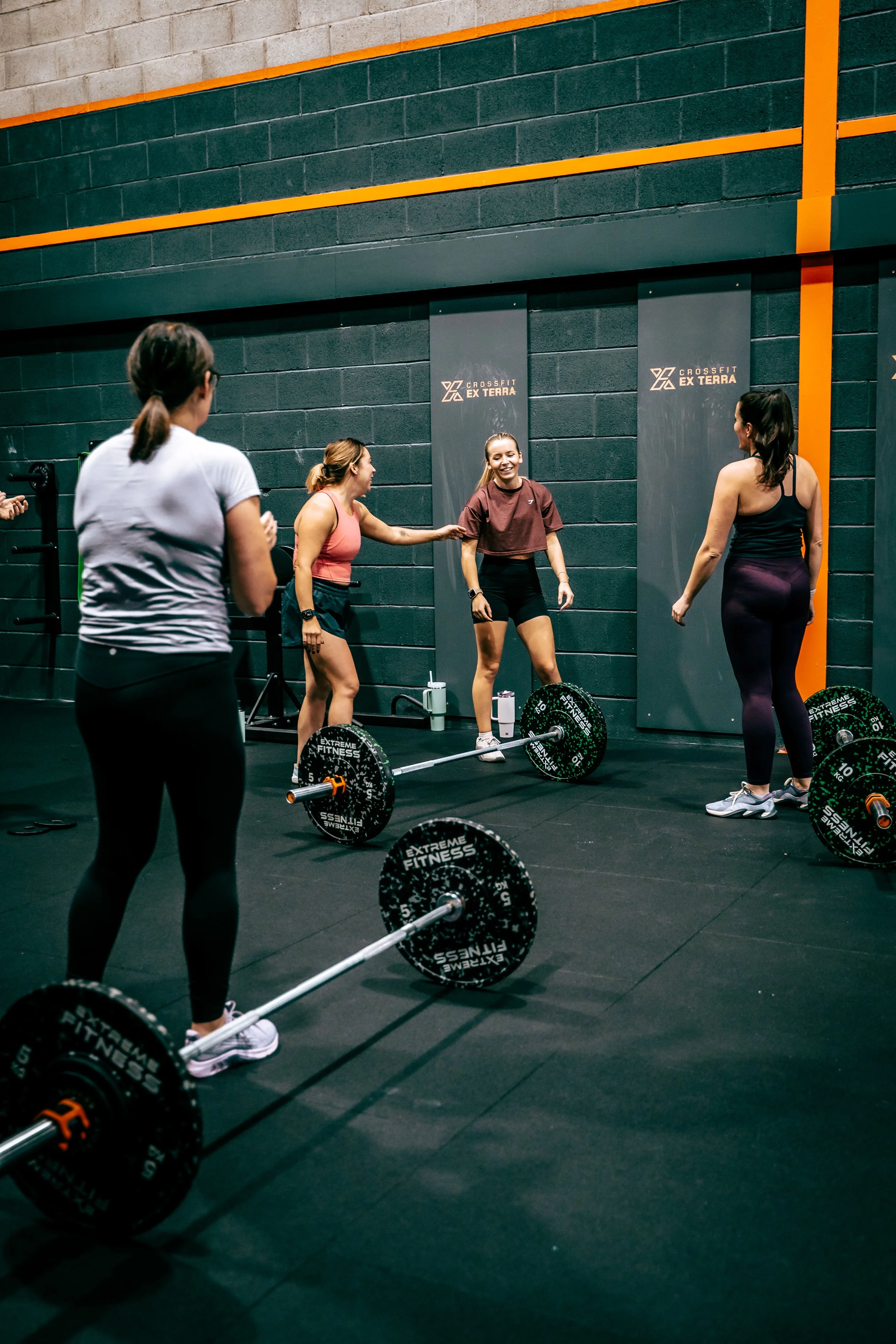 Four women preparing for a CrossFit workout in a gym with black walls, featuring multiple barbells and weights on the floor.