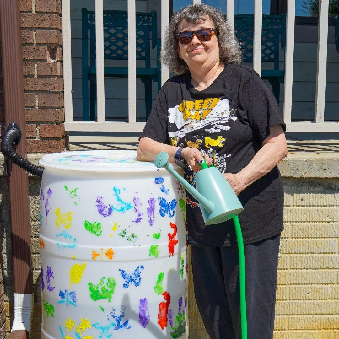 A woman with gray curly hair wearing sunglasses, standing outside next to a colorful decorated barrel and holding a watering can and gardening tools.