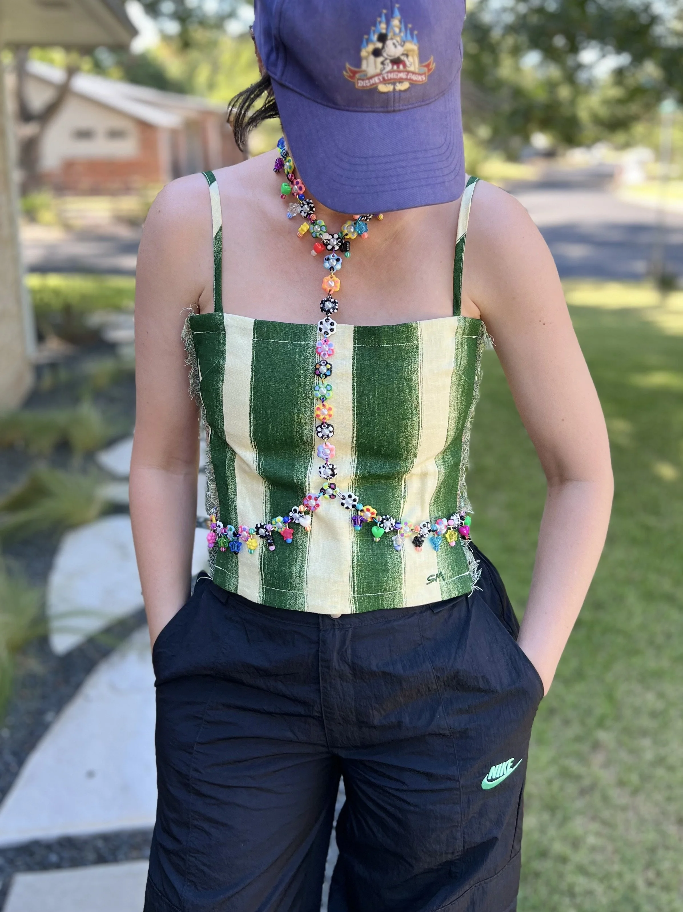 Person wearing a blue Disney theme park cap, a colorful beaded necklace, a green and beige striped sleeveless top, and black Nike pants, standing outdoors on a lawn with trees and houses in the background.