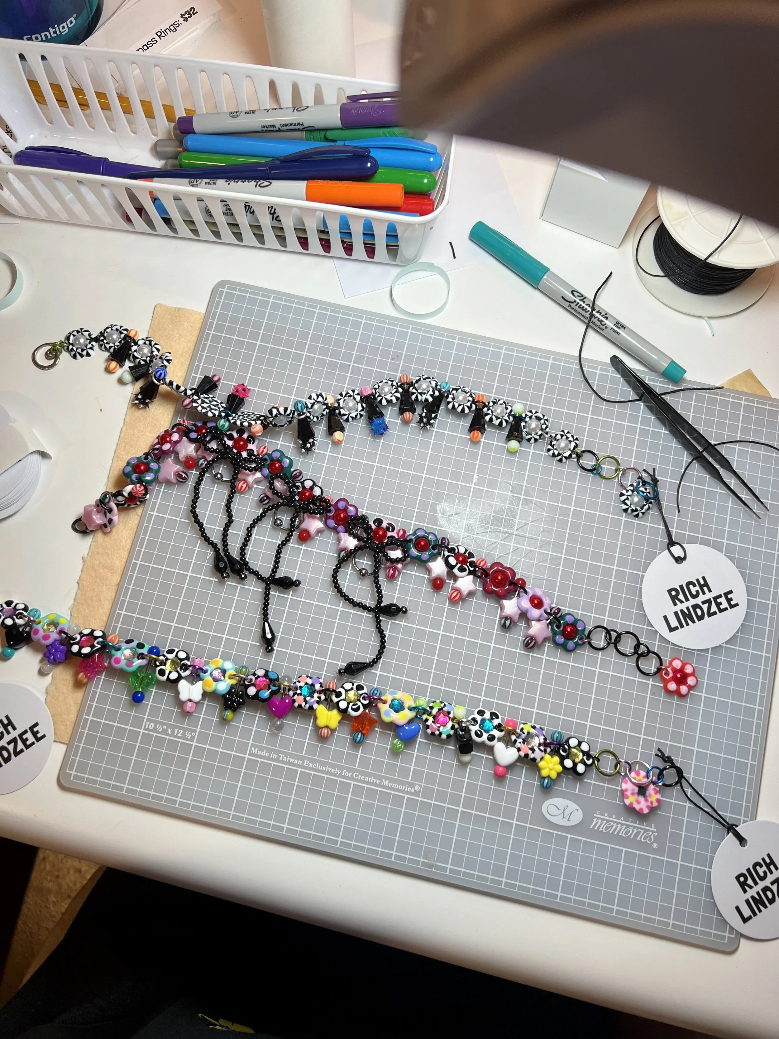Colorful beaded necklaces with various shapes and charms on a crafting mat, surrounded by markers, scissors, and crafting supplies.
