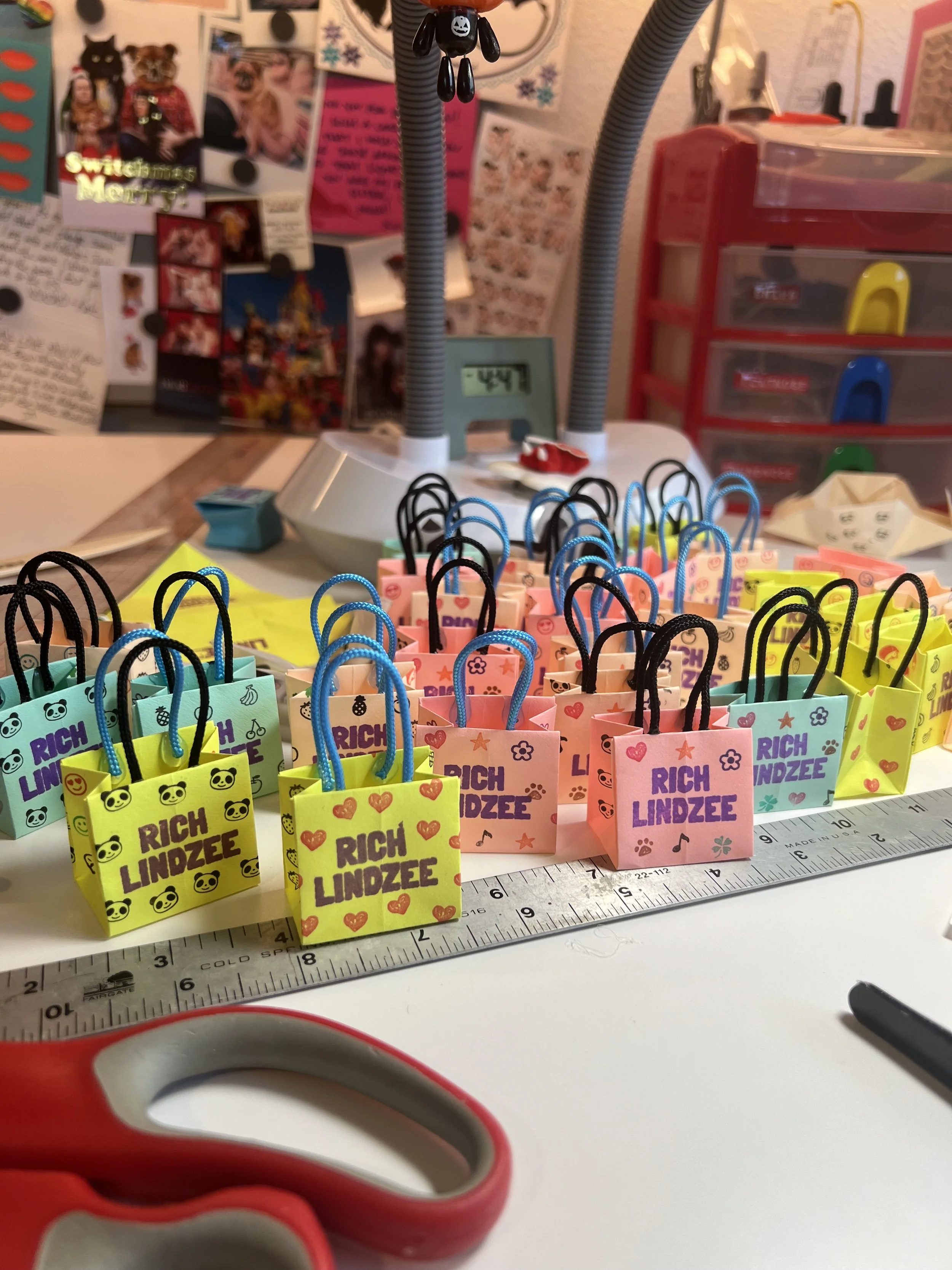 Colorful small gift bags with handles, labeled 'Rich Lindzee', arranged on a table with a ruler, scissors, and various craft supplies in the background.