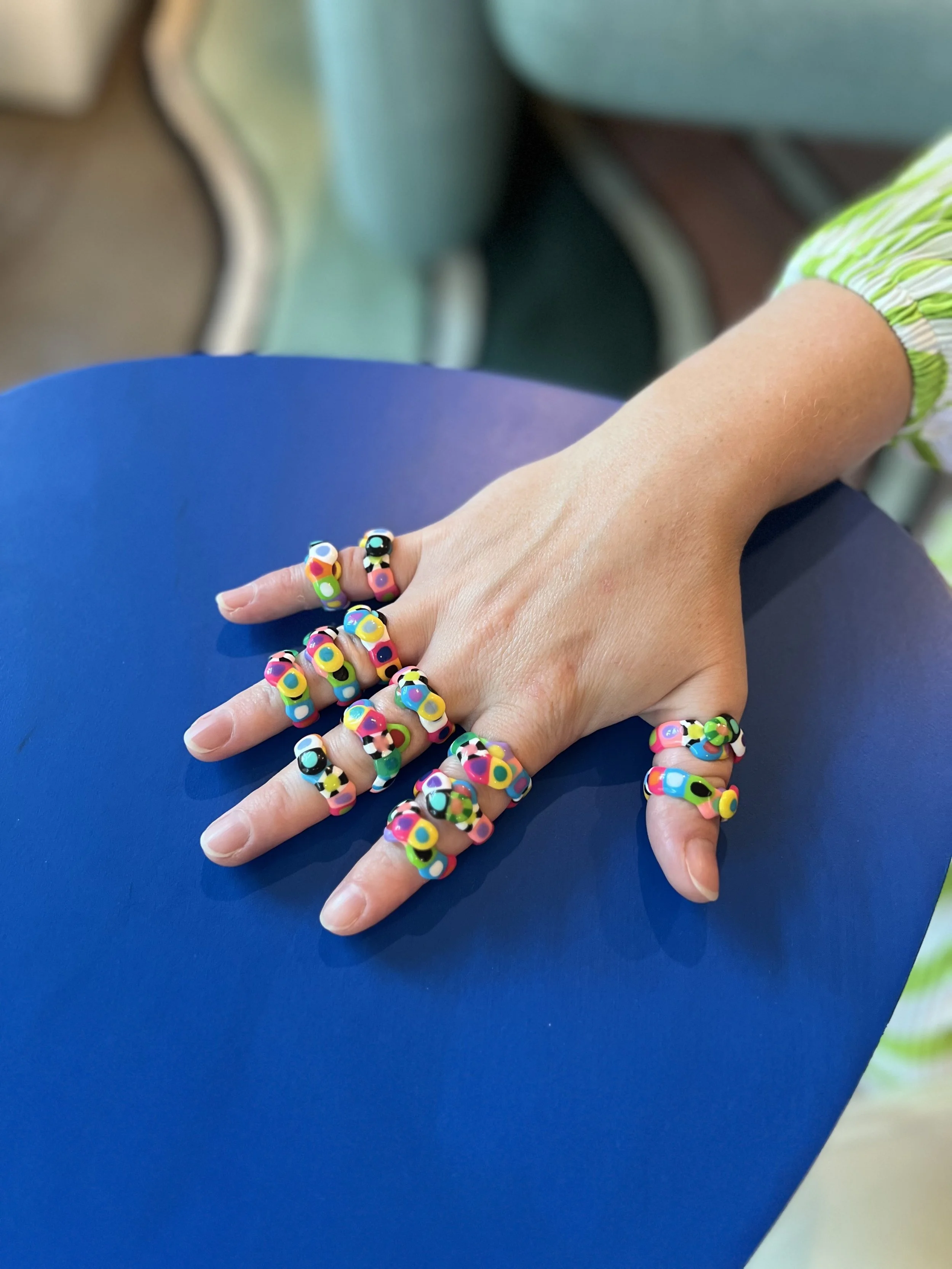 A person's hand resting on a blue surface, adorned with multiple colorful rings with large, eye-catching polka dot and abstract designs.