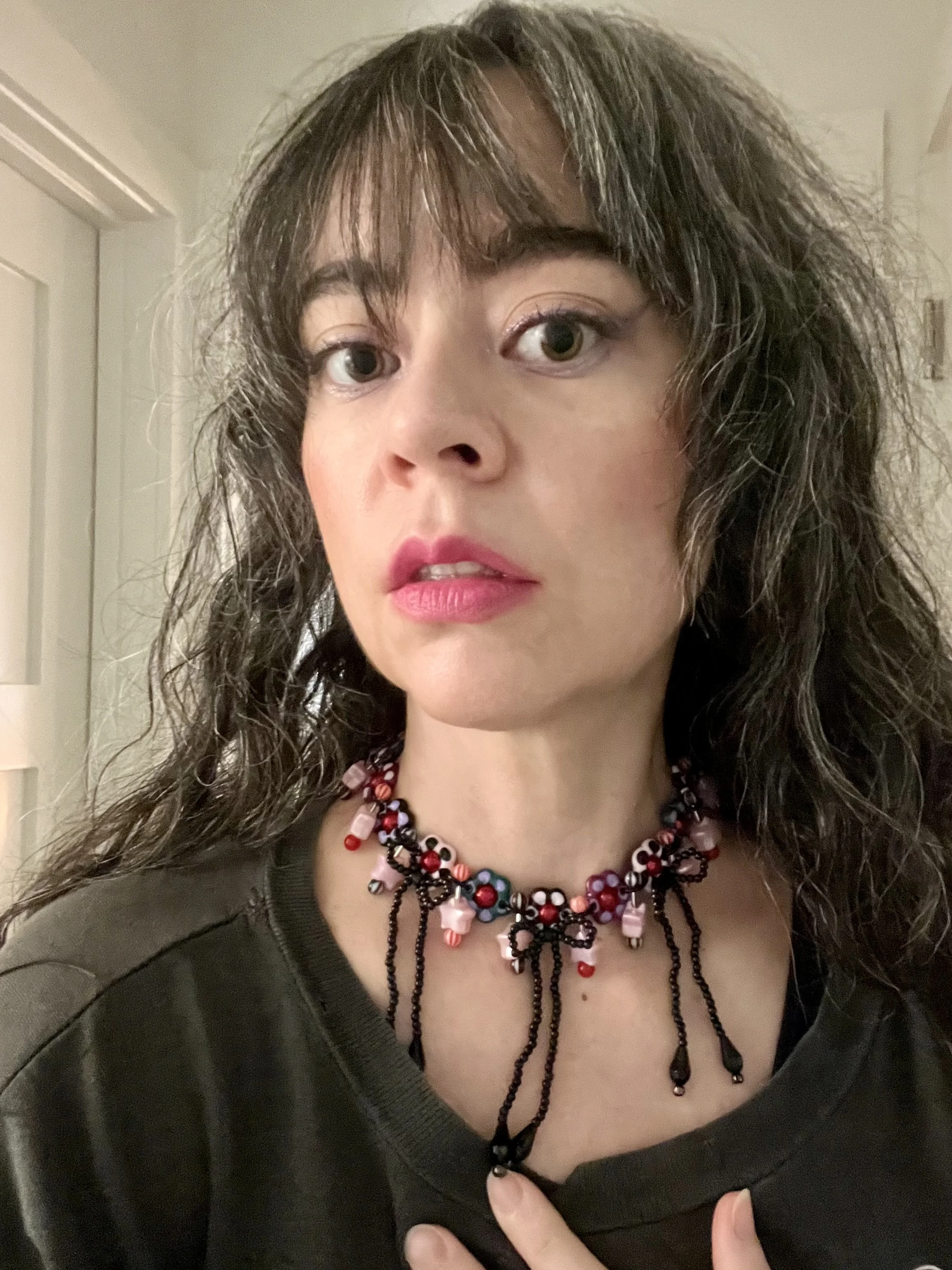A woman with curly, dark hair and fair complexion wearing pink lipstick and a beaded necklace with black, pink, red, white, and blue beads, dressed in a black shirt, standing indoors.
