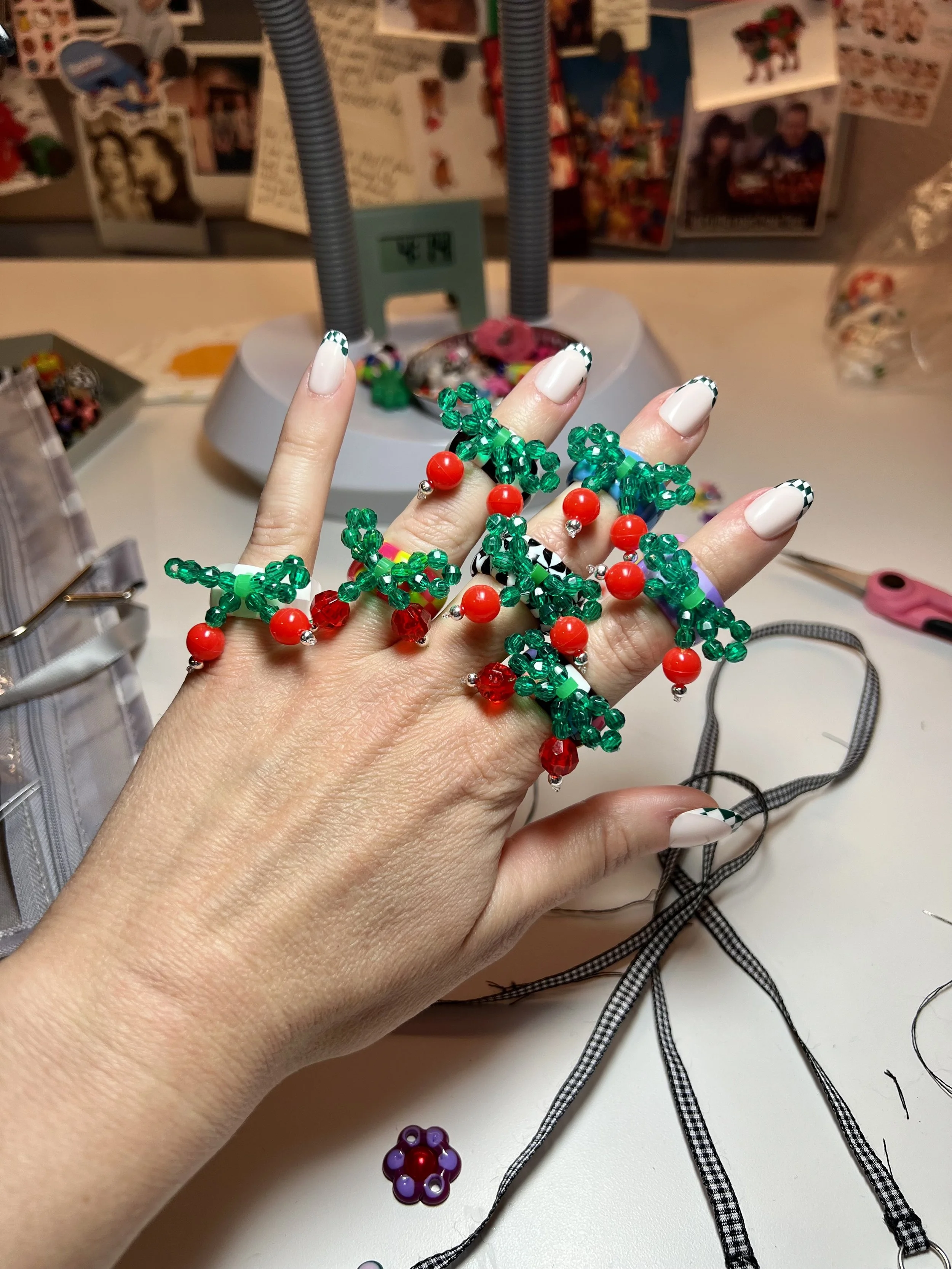 Hand with fingers extended, decorated with multiple Christmas-themed bead bracelets featuring red, green, and black and white beads, on a worktable with jewelry-making materials and a tissue box in the background.