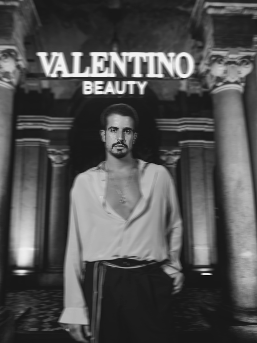 Cartaz publicitário preto e branco de Valentino Beauty com uma pessoa de roupas casuais no centro, em um cenário com colunas clássicas.