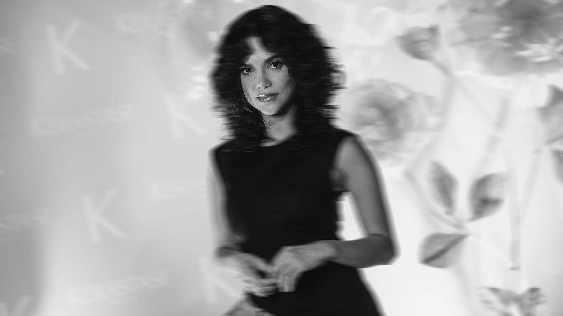 Mulher com cabelo cacheado e vestido preto, em um fundo desfocado com elementos de publicidade ou decoração