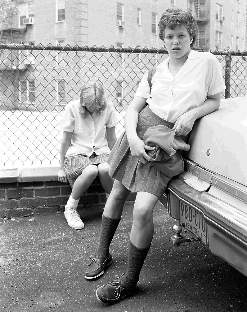 Two schoolgirls in gym uniforms, one leaning against a car and the other sitting on a brick ledge by a chain-link fence in an urban setting.