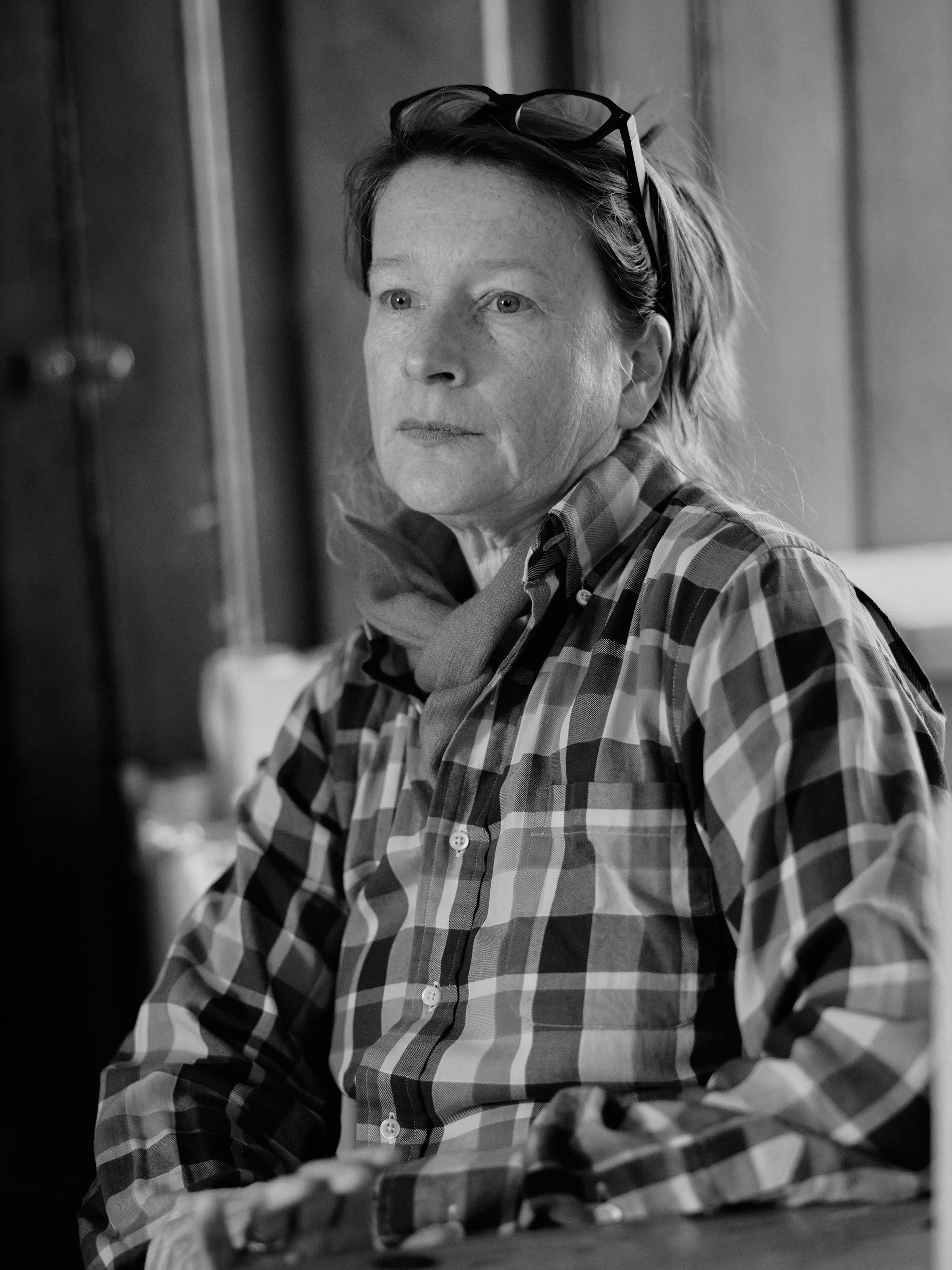 A black and white photo of a woman with short hair, wearing glasses on her head, a checkered shirt, and a sweater. She is looking to the side with a serious expression.