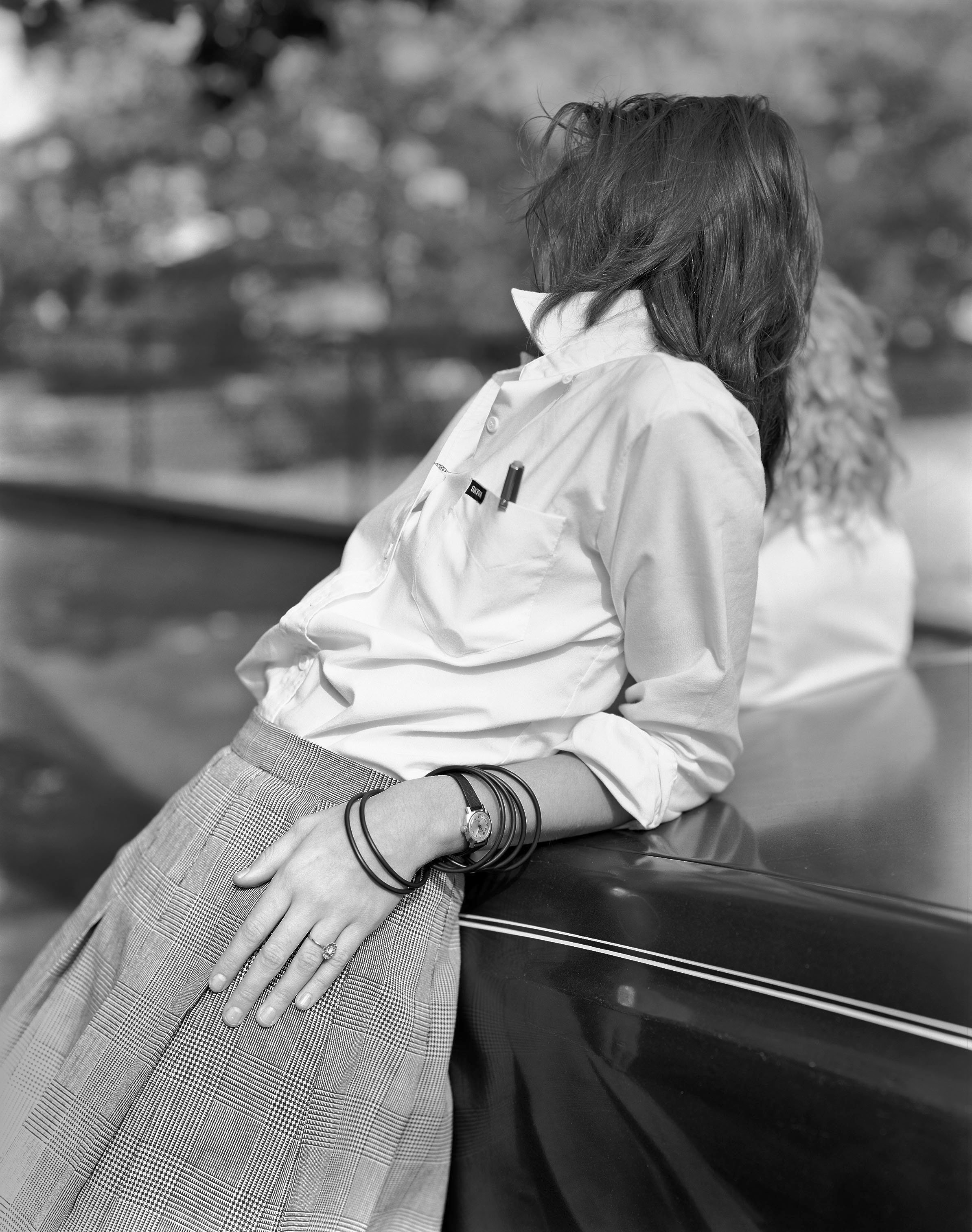 A woman with long hair sitting on a black surface, wearing a white shirt and patterned pants, with her hand resting on her lap and wearing a watch and bracelets. Another person is partially visible in the background.