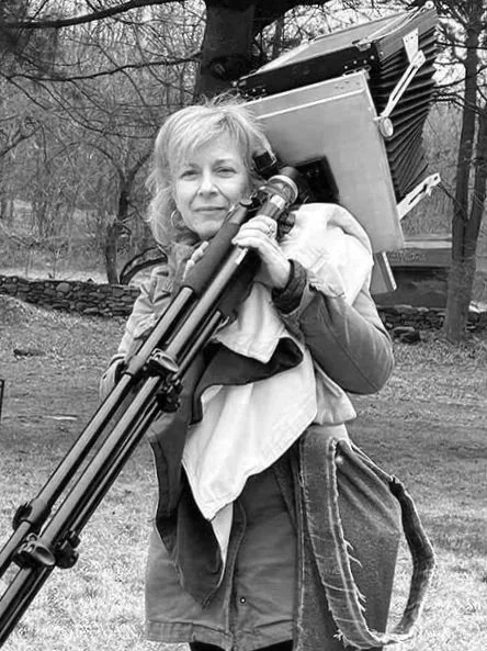 A woman outdoors carrying a large camera and tripod on her shoulder.