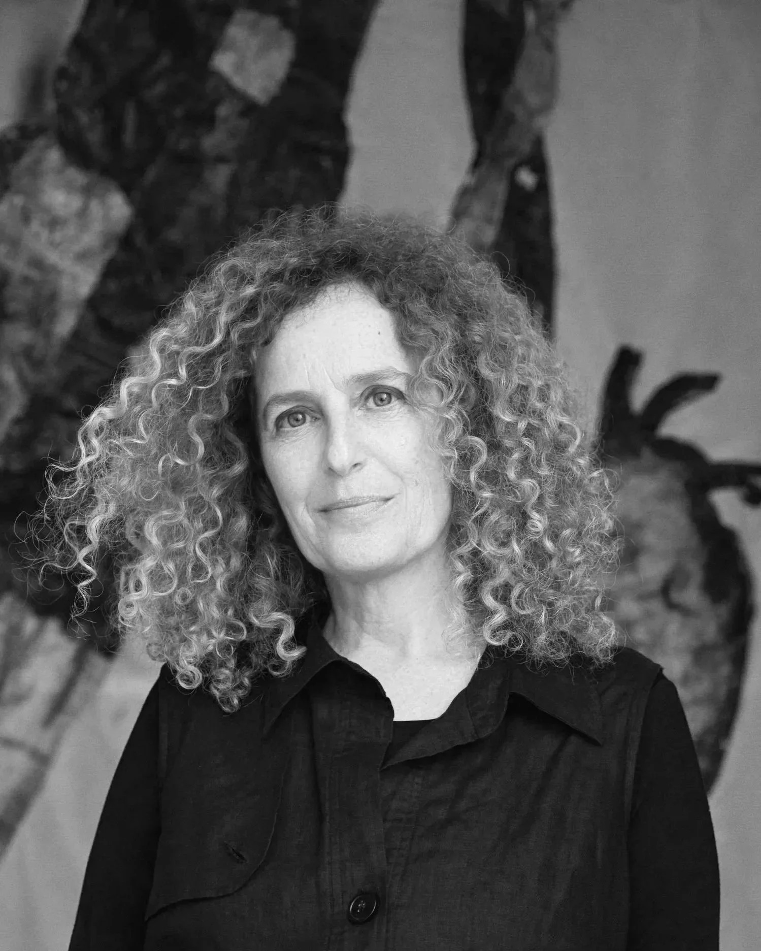 A woman with curly hair wearing a dark blazer and a large button, posing in front of a piece of abstract art, looking directly at the camera.