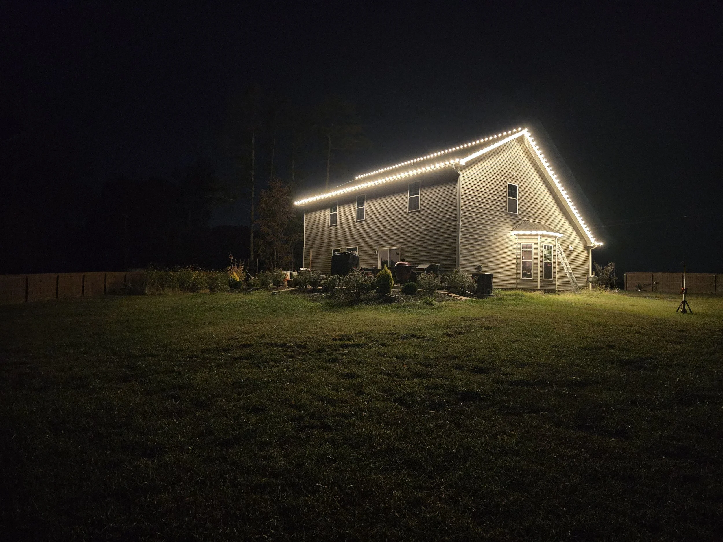 A two-story house at night with exterior lights outlining the roof and eaves. The house has gray siding, several windows, a back door, and a ladder leaning against the side. The house is surrounded by a grassy yard and a wooden fence in the backgroun