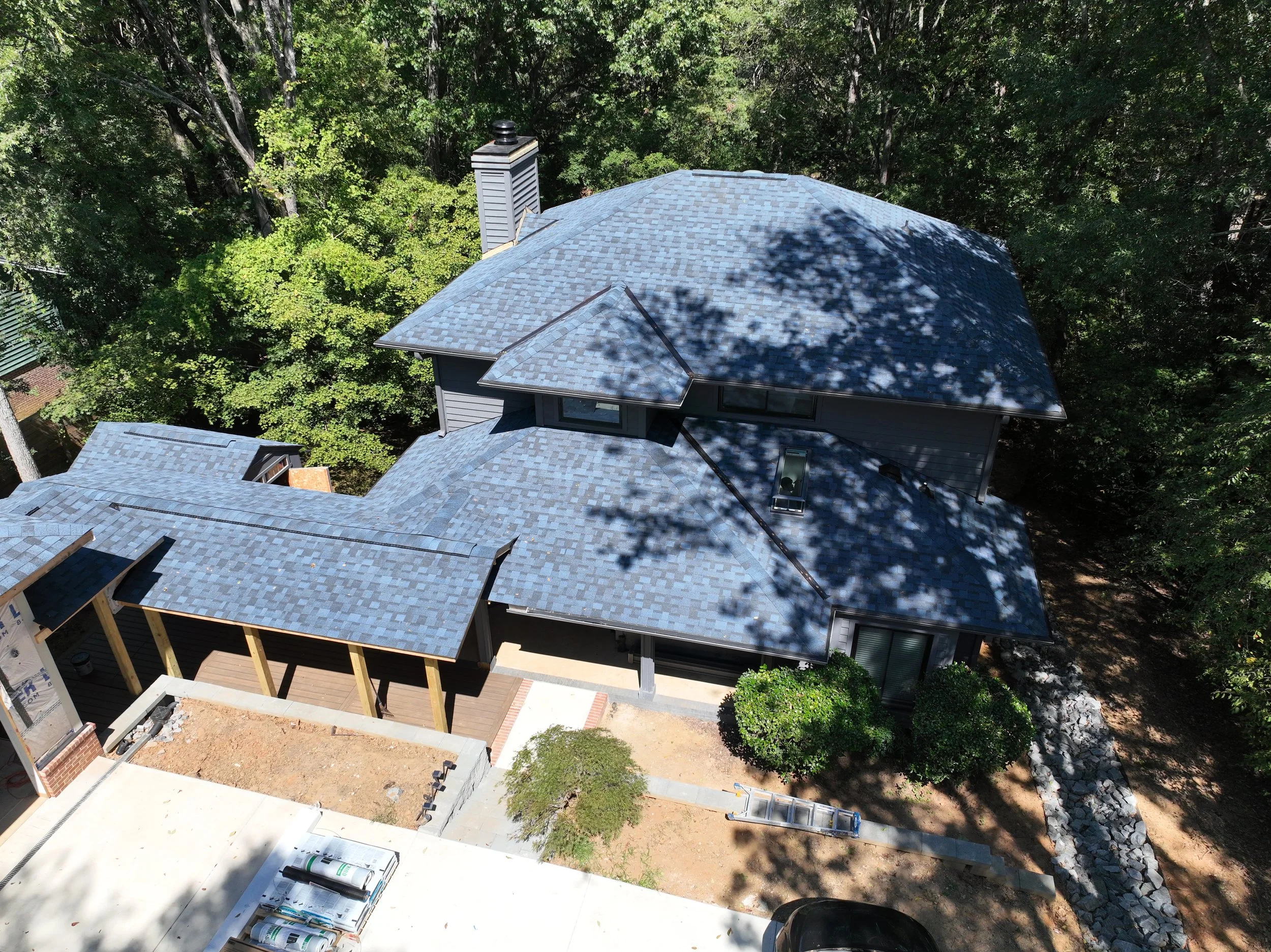 Aerial view of a house under construction with a blue-gray shingled roof, surrounded by trees. There is a wooden porch on one side, and construction materials visible nearby.