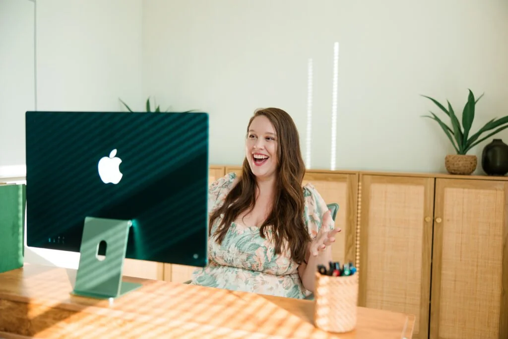 A woman sitting at a wooden desk, smiling and gesturing towards a computer with an Apple logo, in a bright room with a light green wall and wooden cabinet with plants in the background.