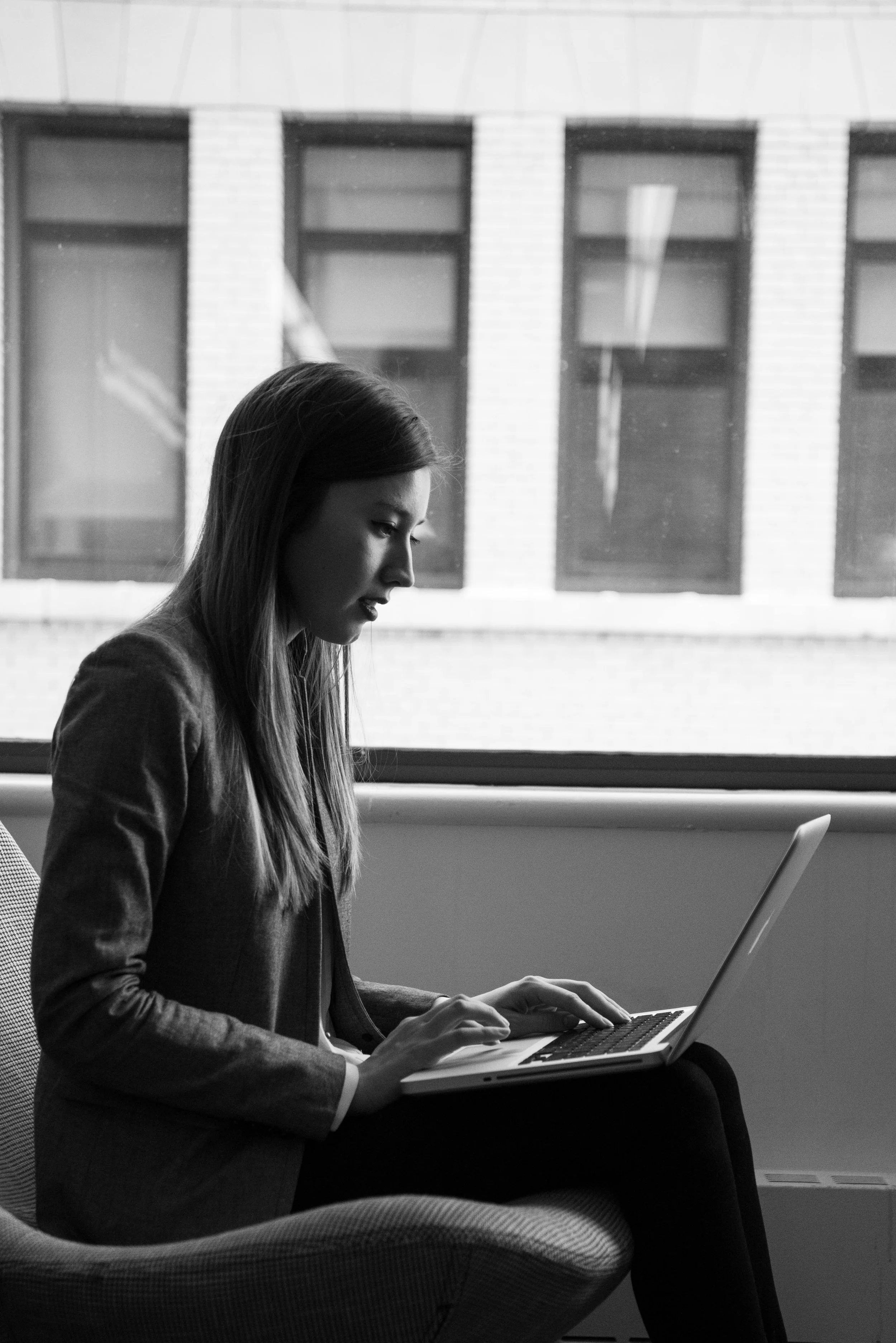 A woman with long hair in a blazer sitting on a chair, using a laptop in front of a large window with a view of a building's brick wall and windows outside.