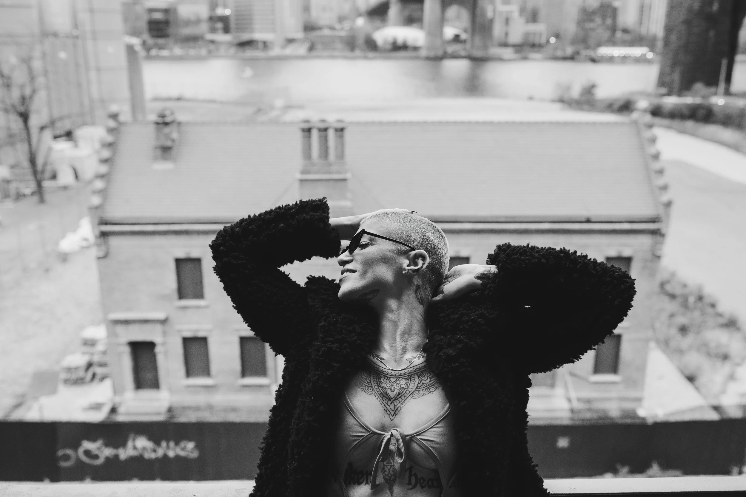 A person with a shaved head, wearing glasses and a furry jacket, poses with arms behind their head on a rooftop. They are smiling and have tattoos visible on their chest and neck. The background includes a cityscape with buildings and a body of water.