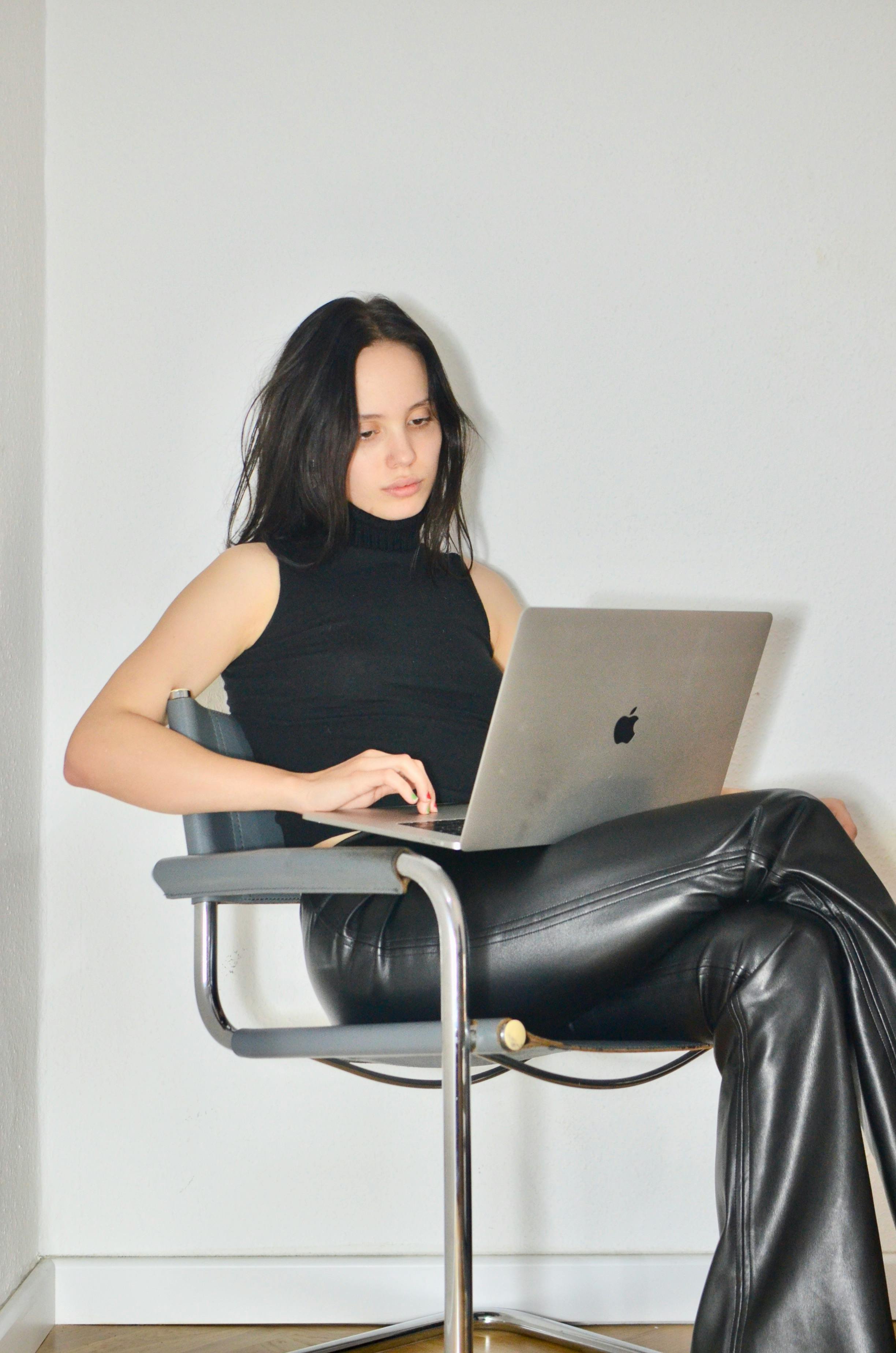 A young woman with black hair, wearing a black sleeveless turtleneck top and black leather pants, sitting in a gray chair with her legs crossed, working on a silver MacBook laptop against a plain white wall.