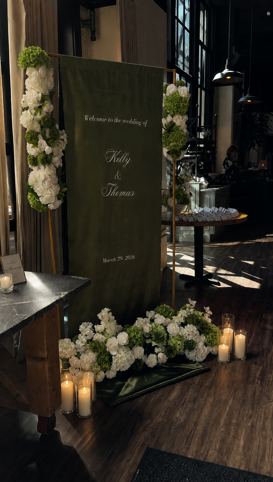A wedding welcome sign decorated with white roses and green hydrangeas, with candles placed at the base, welcoming guests to Kelly and Thomas's wedding on March 29, 2026, inside a dimly lit venue with large windows.